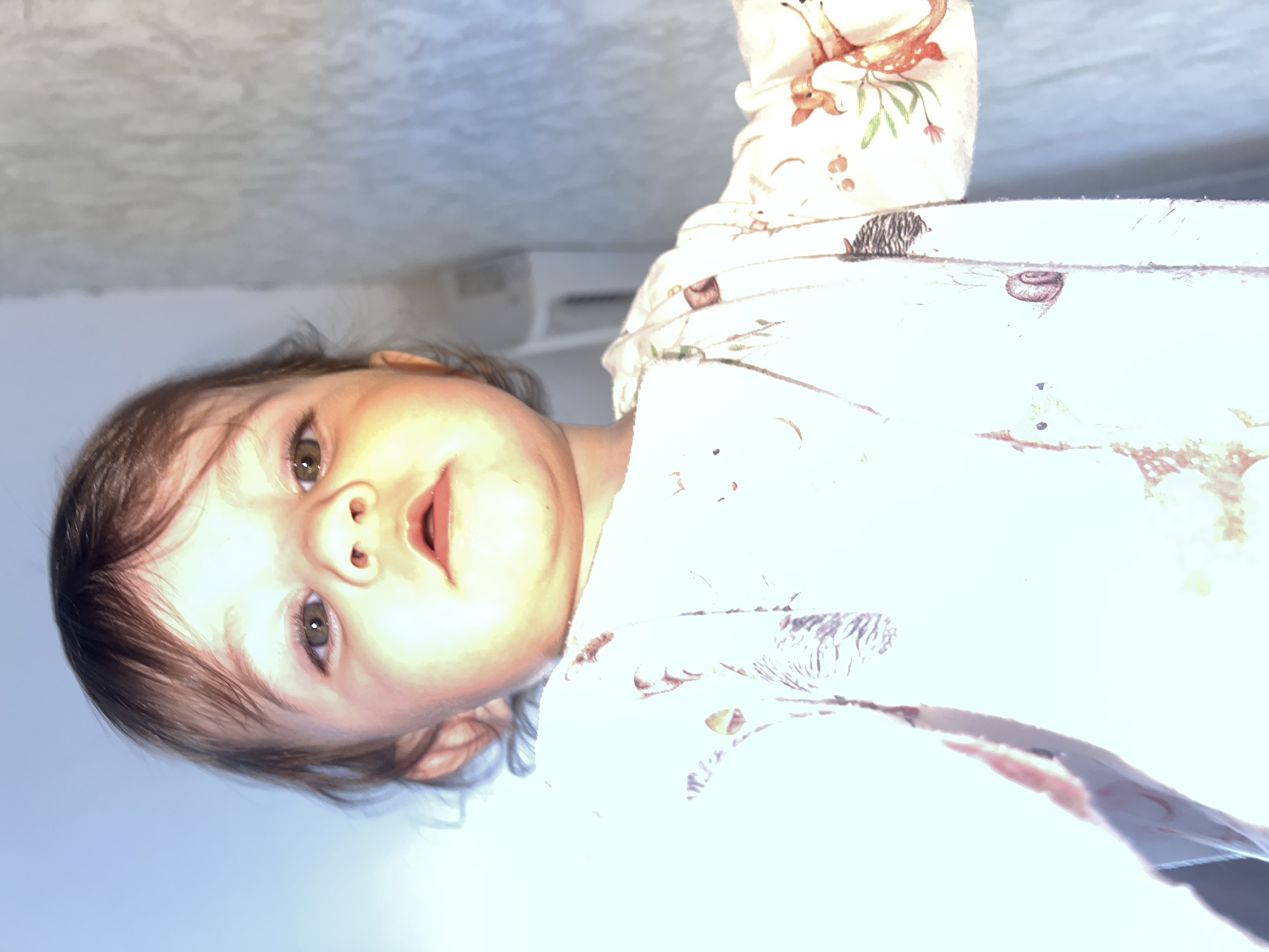 A young child with brown hair and eyes, wearing a white garment with a woodland animal print, is photographed from a low angle, making the child appear to look down at the viewer. The background appears to be a ceiling and wall. There is no identifiable information to indicate a specific location in the city of Elena, Bulgaria.