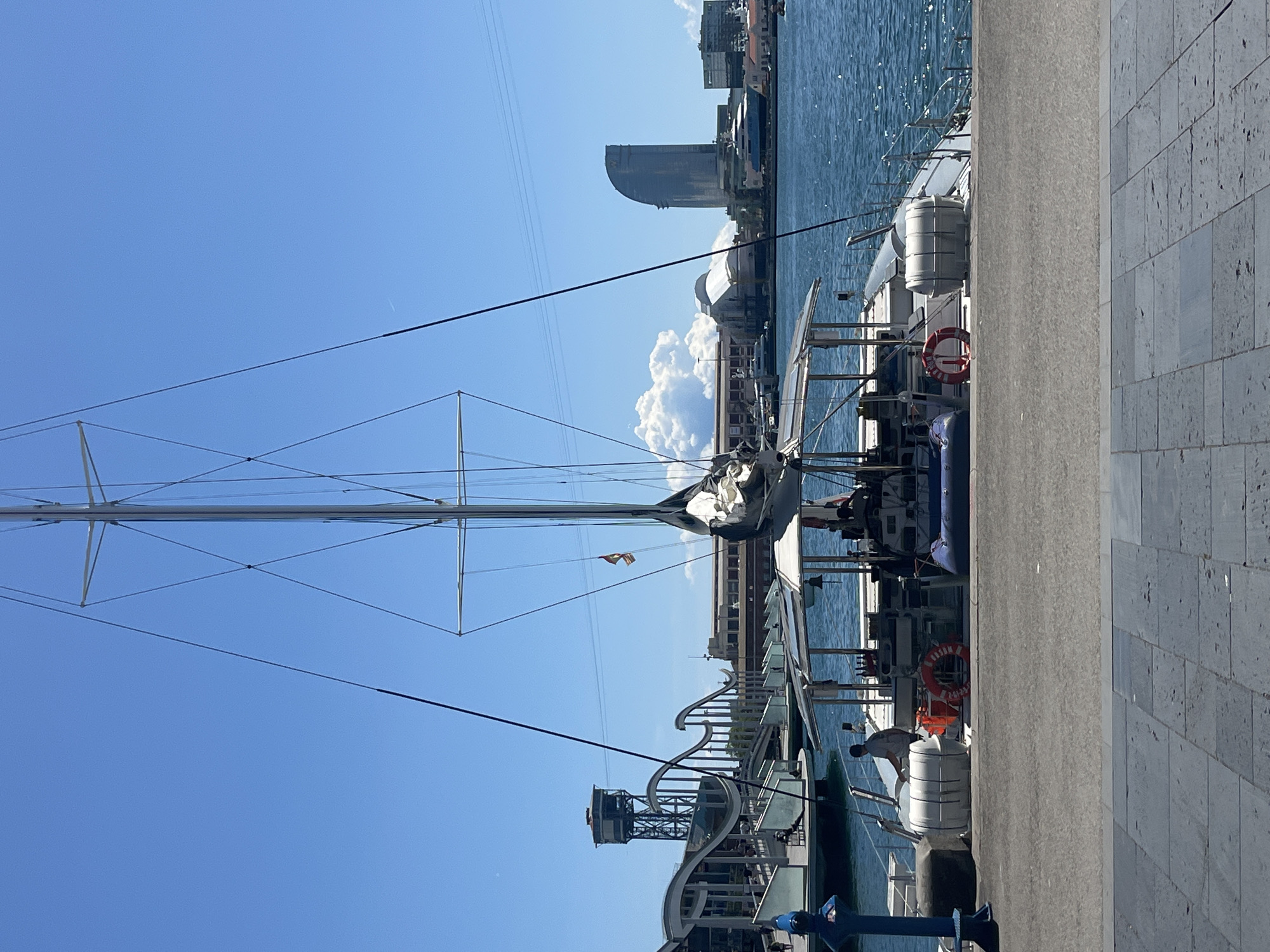A yacht is docked at a pier in Barcelona, Spain. The yacht's mast extends high into a blue sky dotted with a large, fluffy white cloud. The Hotel Arts Barcelona is visible in the distance. A man in a light-colored shirt is standing on the pier next to the yacht, looking down at something. A red life preserver is visible on the yacht. The pier is made of gray stone slabs. A building with a lattice-like structure is also visible near the pier.