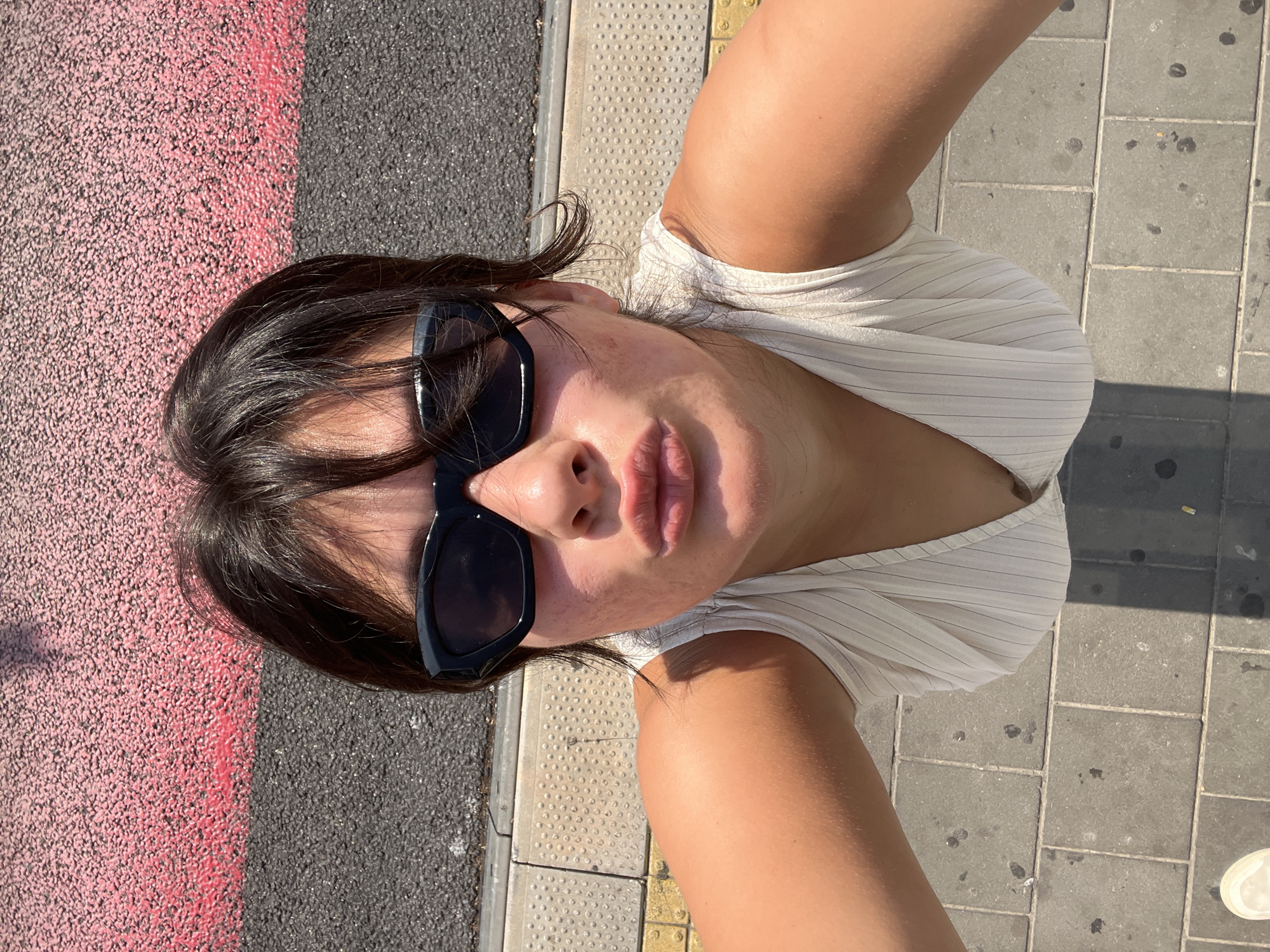A young woman is taking a selfie outdoors in what appears to be an urban setting. She is wearing black sunglasses and a white ribbed top, and is puckering her lips. Her arms and shoulders are visible, as is her dark hair. The background shows a textured surface, possibly pavement or a wall, with distinct pink and gray sections. The ground beneath her is paved with square tiles. The lighting suggests it is daytime. There is no specific indication of Kiryat Motzkin, Israel, in the image itself.