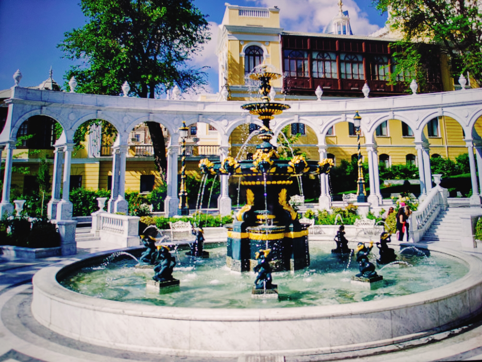 The image shows a large, ornate fountain in the foreground, with several smaller figures of children playing in the water. The fountain is decorated with gold accents and appears to be made of dark stone. Behind the fountain is a white, colonnaded walkway with arched openings. In the background, there is a yellow building with a dark wooden balcony and a domed structure. Lush green trees are visible on the left side of the frame. Two women are standing on a staircase on the right side of the image, one wearing a green and yellow patterned outfit and the other in a black dress and a pink head covering. The scene is likely a public square or park. This is likely the Fountain Square in Baku, Azerbaijan, a popular tourist destination known for its fountains and surrounding architecture.