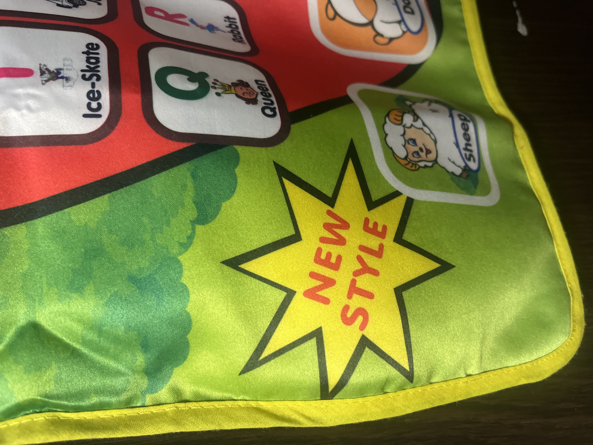 The image shows a close-up of a green fabric with yellow edging, likely part of a child's learning mat or poster. The fabric has a printed design of green clouds in the background. In the foreground, a yellow star shape with a black outline contains the words "NEW STYLE" printed in orange. To the right of the star, there are several other printed squares, each with a letter, an illustration, and a word. These include "Ice-Skate," "Queen," and "Rabbit." Below the "Rabbit" square, there is a square with an illustration of a sheep and the word "Sheep." Another square with an illustration of a dog and the word "Dog" is partially visible at the bottom right. The image does not provide any information or visual cues to suggest its location in the city of Tarnivtsi, Ukraine.