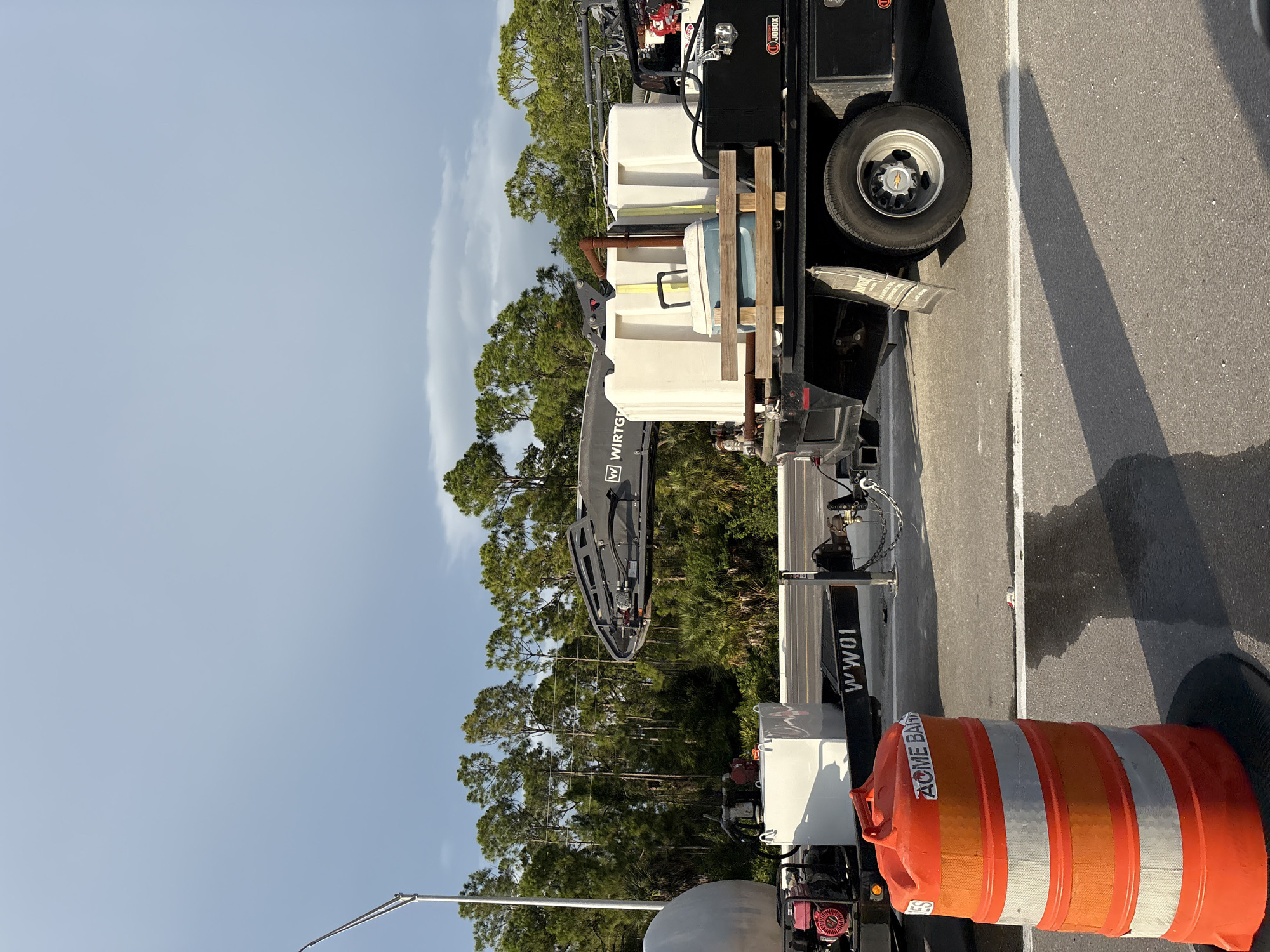 An outdoor, medium shot captures a flatbed truck with multiple water tanks and equipment parked on a road, likely for construction or maintenance work, under a cloudy sky. The truck has "W WIRTS" visible on a piece of equipment, and "WW01" on its side. In the foreground, an orange traffic barrel with reflective bands is positioned on the asphalt. The background features a dense line of tall, green trees. The scene is illuminated by natural light, casting shadows on the road.