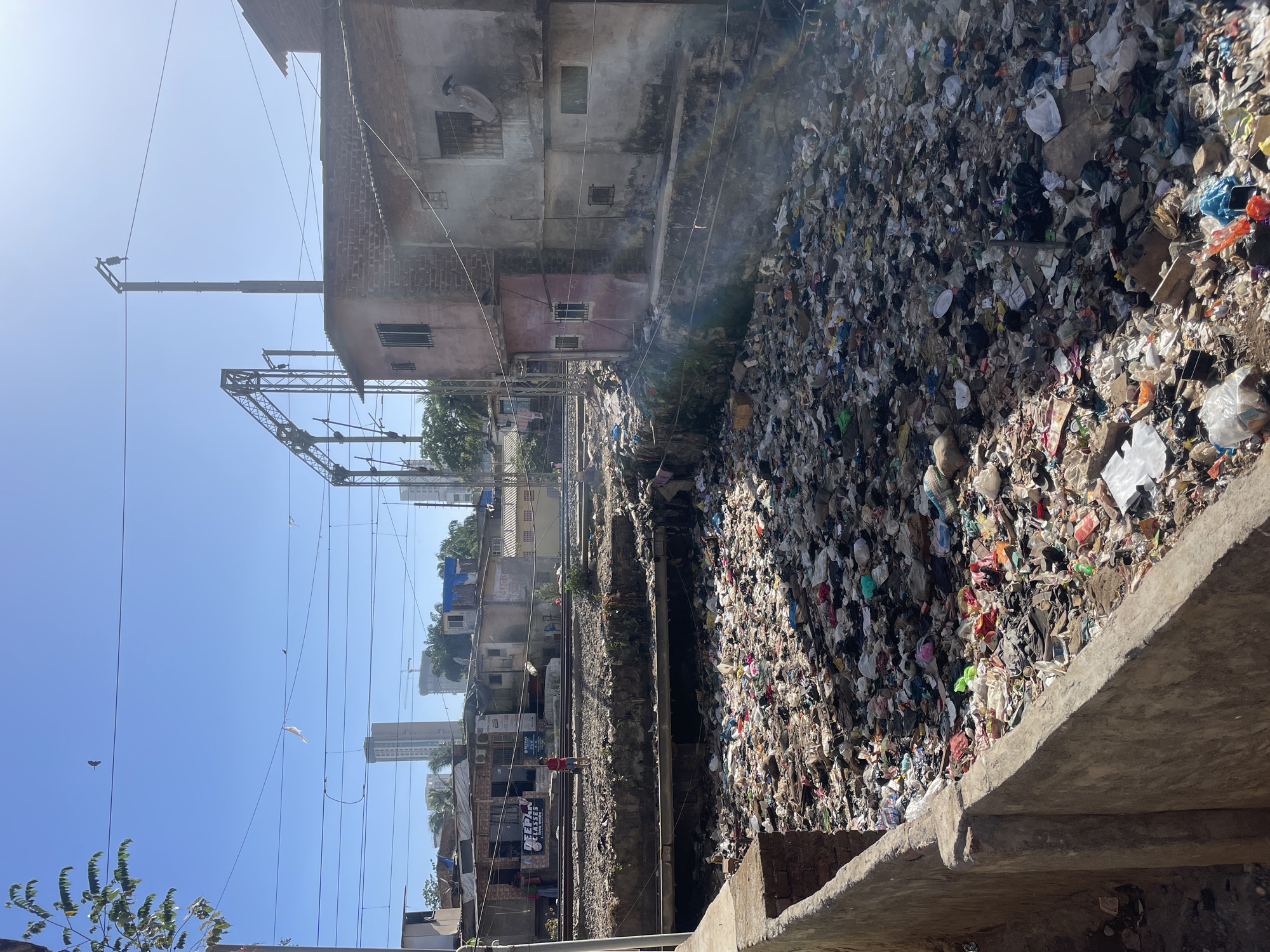 The image depicts a scene in Mumbai, India, characterized by significant environmental pollution. A large open area, seemingly a drainage canal or ravine, is filled with an immense amount of garbage and waste. This extends across the frame and suggests a widespread problem of waste management in the area. Adjacent to the waste-filled area are residential buildings of varied construction and condition, appearing densely packed and indicative of a lower-income area. Railway tracks run parallel to the garbage area, adding to the urban environment. Overhead power lines and supporting structures are visible against the bright sky. A person in a red shirt is seen near the train tracks. The scene presents a stark contrast between the basic infrastructure and the severe environmental conditions, indicative of an urban area struggling with overpopulation and waste disposal.