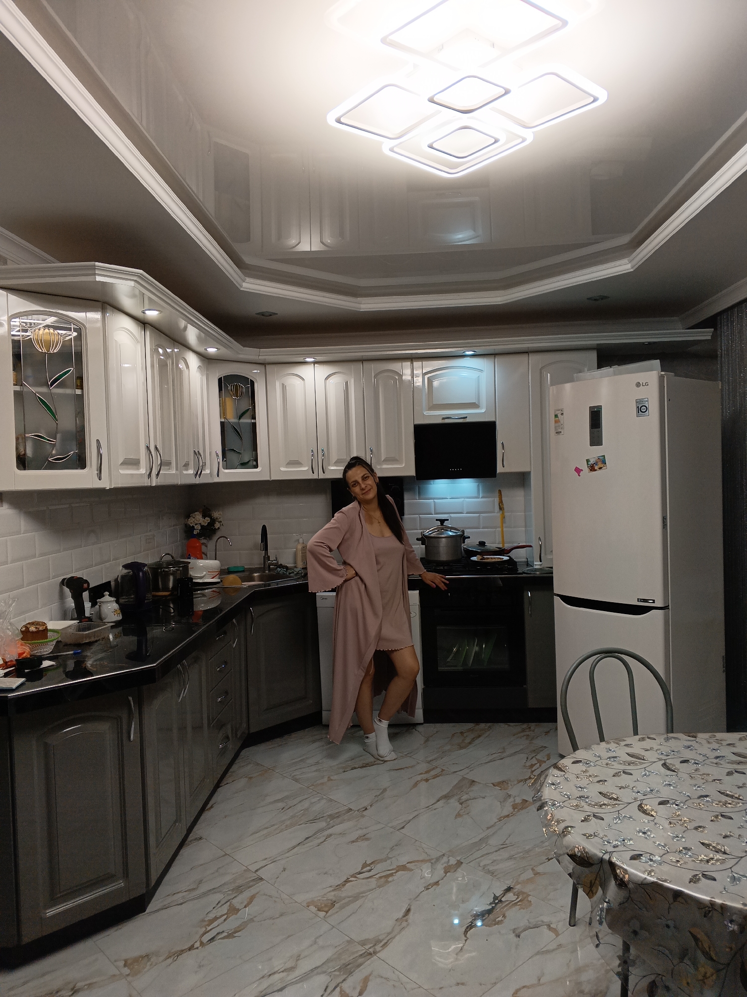 The image shows a modern, well-lit kitchen in Andreyevo, Russia, with a woman standing centrally. She has dark hair, wears a light pink, long-sleeved cardigan over a shorter garment, and white socks. She poses casually, with her left hand on her hip and her right arm resting on the black stove, looking towards the camera with a gentle smile.
The kitchen features white upper cabinets, some with glass inserts displaying stained-glass-style floral patterns, and darker grey lower cabinets. Black countertops and a white subway tile backsplash complete the workspace. Visible appliances include a white LG refrigerator on the right, which has the text "LG INVERTER COMPRESSOR" and a "10 YEAR WARRANTY" logo. A black range hood is positioned above a black stove and oven. Various items are on the countertops, such as a black electric kettle, a drill, a white teapot, and a baked good.
The ceiling is a glossy white stretch ceiling with decorative crown molding and a prominent multi-square LED light fixture, which is reflected in the surface. The floor is tiled with a light grey and brown marble-effect pattern. In the foreground right, a round table covered with a floral-patterned tablecloth and a grey metal chair are partially visible. The scene depicts a domestic setting, likely under artificial lighting during the evening.