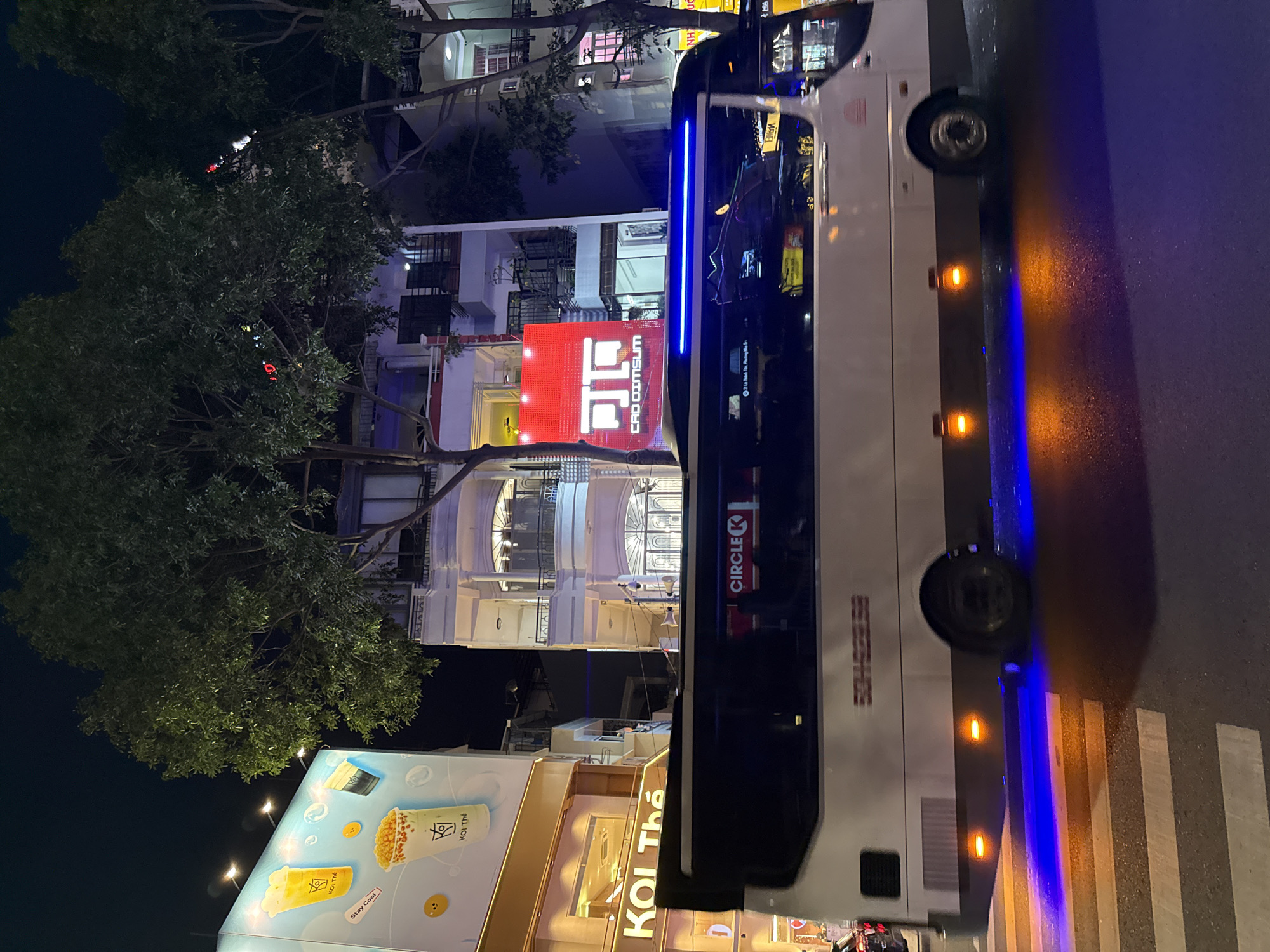 This is a nighttime, eye-level, full shot of a street scene in Nha Trang, Vietnam. A white bus with blue accent lighting underneath is driving across a crosswalk in the foreground. The bus has the Circle K logo on its side. In the background, there are buildings with lit windows and signs. A large, brightly lit red sign with white text reading "JIN CAO DIMSUM" is visible. The street is wet, reflecting the lights. Trees with full foliage line the street.