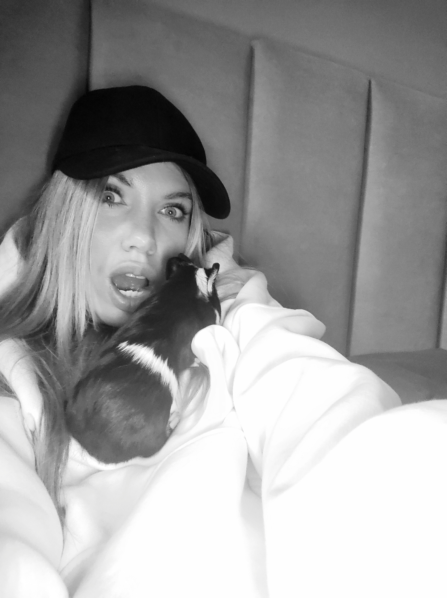 A black and white, close-up photograph captures a woman and a small pet indoors, likely in Votkinsk, Russia. The woman, positioned centrally, has long, light-colored hair and is wearing a black baseball cap with a brim and a light-toned (likely white) hooded garment. Her expression is one of wide-eyed surprise or playful shock, with her mouth open and her gaze directed towards the viewer.
She is holding a small, black and white guinea pig or similar rodent close to her face, positioned on her right side. The animal features distinctive white markings against its dark fur, particularly on its back and side. This interaction creates an intimate and candid moment.
The background shows a padded or upholstered wall, possibly a headboard, composed of vertical rectangular panels in shades of light grey. The lighting indicates an indoor setting. No text is visible in the image. The photograph focuses on the expressive interaction between the woman and her pet.