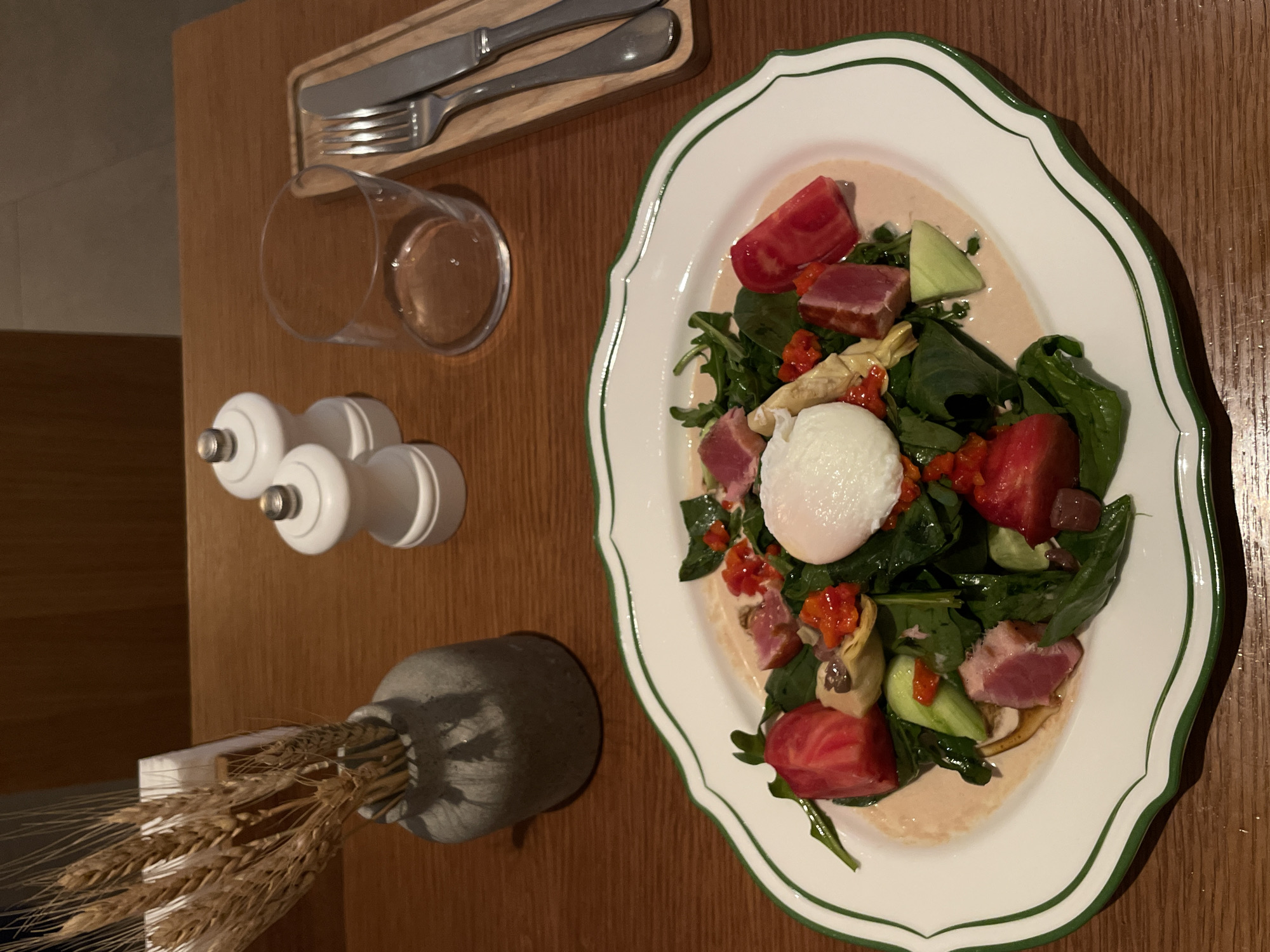 The image captures an overhead view of a table setting, likely in a restaurant, with a salad prominently displayed on a decorative white plate edged with a green line. The salad consists of a poached egg, leafy greens, cubed red tomatoes, and artichoke hearts, all drizzled with a light sauce. To the left of the plate are dining utensils (knife and fork) on a small wooden tray, a glass partially filled with liquid, and a set of white salt and pepper shakers. A small grey vase containing wheat stalks is also present. The table has a wooden surface. The environment suggests an indoor dining area, possibly a restaurant in Moscow, Russia.