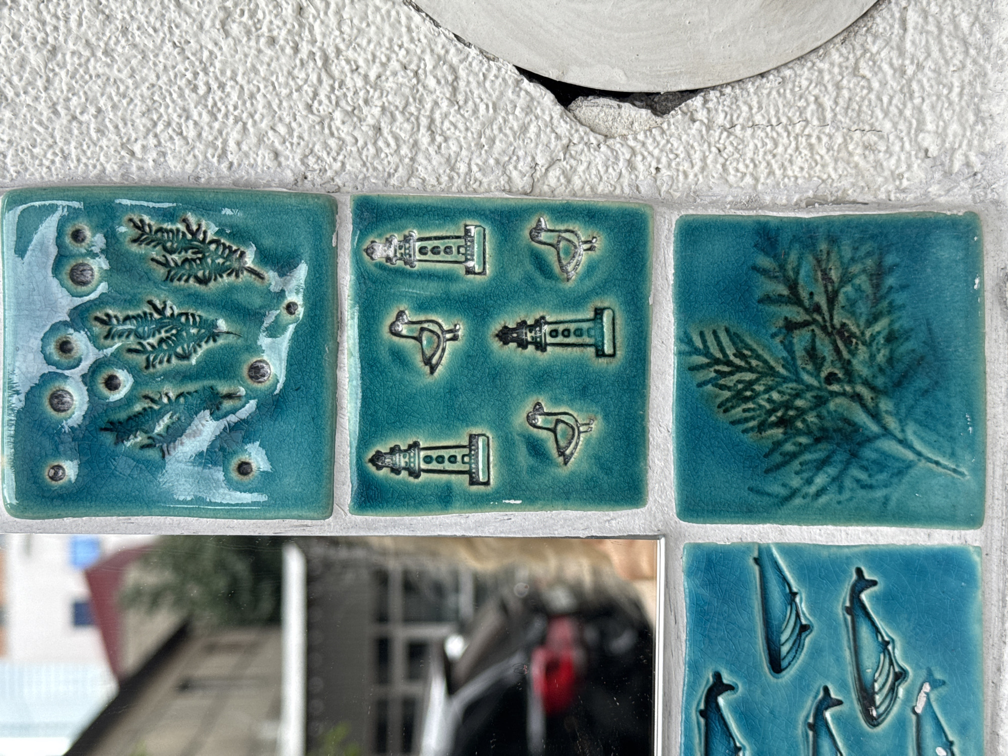 The image shows a close-up of decorative ceramic tiles mounted on a white textured wall. The tiles are predominantly teal and depict various marine-themed designs. In the top tile, there are impressions of green foliage and small circles, possibly representing bubbles. The second row features three lighthouse motifs and two bird illustrations, all in outline. The bottom left tile displays a pattern of several whales swimming. To the right of the whale tile is a tile with a detailed illustration of green foliage. The tiles appear to be part of an exterior decorative element. A mirror is positioned to the left of the tiles, reflecting a blurred image of buildings, trees, and a dark-colored car. This scene is located in the city of Batumi, Georgia.