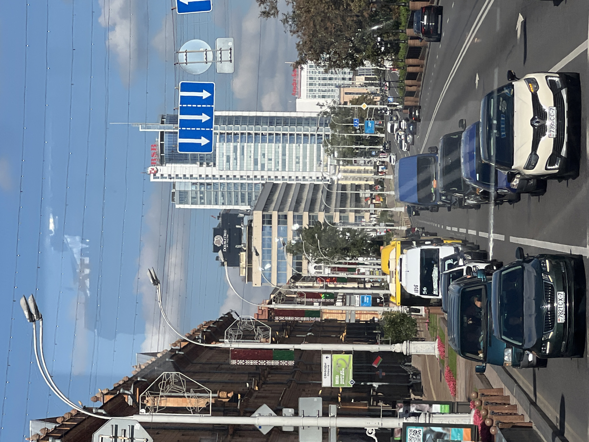 A forward-facing, slightly elevated shot captures a city street in Minsk, Belarus, on a sunny day with a blue sky dotted with white clouds. The street is filled with traffic, primarily cars and vans, moving in the same direction. In the foreground, a dark green Skoda sedan with license plate 7821CX-7 is the closest vehicle. Behind it are several other vehicles, including a white Renault SUV with license plate 7395CC-7, a blue van, and a dark-colored van.
The street is lined with buildings on the left and trees and a bridge on the right. The buildings on the left are a mix of older, ornate architecture and modern high-rises. One building has flags of Belarus hanging from lampposts. Traffic lights and street signs are visible along the road. In the background, a tall, modern skyscraper with the letters "RSB" prominently displayed in red can be seen. Further in the distance, other tall buildings, including one with "Alifa-Bank" written in red, contribute to the urban skyline. The street is paved and has lane markings. The overall scene depicts a busy urban environment.