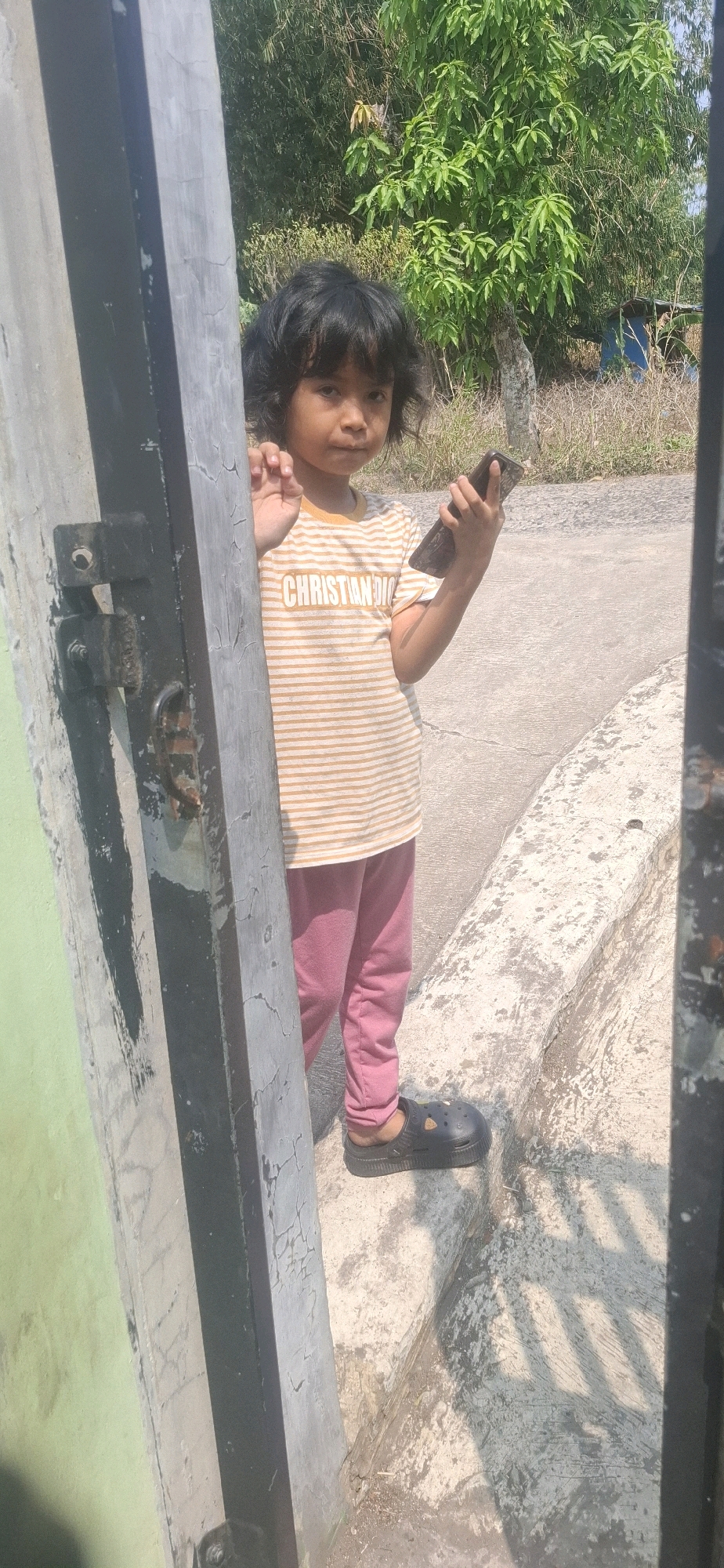 In Cibinong Hilir, Indonesia, a young child stands near an open doorway. The child, with dark, curly hair, is wearing a horizontally striped yellow and white t-shirt with the text "CHRISTIAN DIO" printed on it, and pink pants. They are wearing black slip-on shoes. The child is holding a dark-colored smartphone in their right hand and is looking towards the camera. The background shows dry vegetation and trees, with a concrete curb and ground. The doorway is framed by weathered gray and green painted posts, with a visible metal latch on the left post. Sunlight casts shadows across the ground.