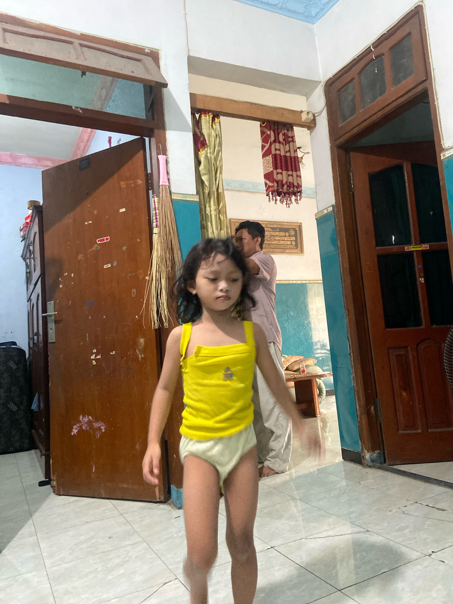 The image depicts an indoor scene in Gelam, Indonesia. In the foreground, a young child wearing a yellow tank top and light-colored briefs stands in the center of the frame. The child has dark, curly hair and is looking down with a neutral expression. Behind the child, a man in a pinkish-purple shirt and light-colored pants is partially visible, appearing to be walking away or looking towards the left of the frame.
The setting appears to be a room with wooden doors and window frames. A large, dark brown wooden door stands to the left, slightly ajar, revealing a dark interior and a broom with a pink handle leaning against it. To the right, another dark wooden door with glass panes is visible. The walls are painted in a light color, with a band of turquoise paint along the lower section. A decorative curtain with a red and maroon pattern hangs in the background, above what appears to be a framed piece of artwork or text on the wall. The floor is tiled with light-colored squares.
The lighting suggests it is daytime, though the specific time of day is not discernible. The overall impression is that of a casual domestic environment. The activity seems to be people moving within a house.