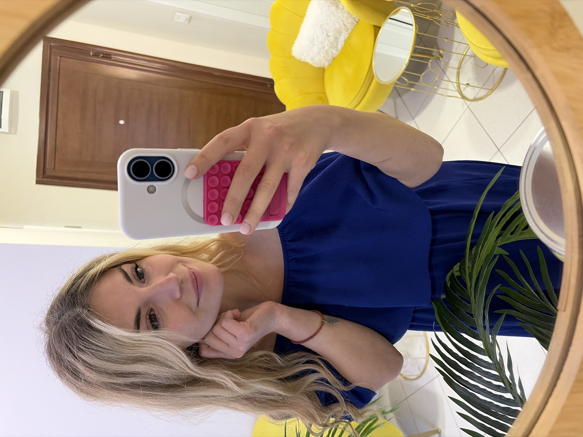 A blonde woman is reflected in a circular mirror, seemingly taking a selfie with a white and pink smartphone. She is wearing a blue dress. The background includes a closed wooden door, a yellow chair with a white cushion, and a small round table. The location is potentially an interior setting in Budva, Montenegro, but this cannot be verified through the image alone.
