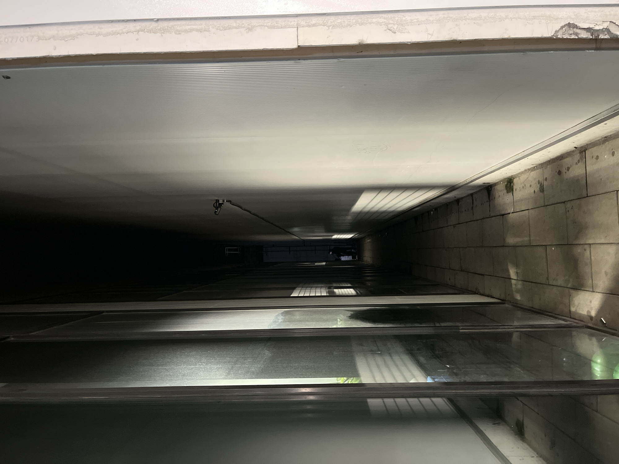 The vertical image presents a deep, narrow shaft viewed from an elevated position within a building in Shumen, Bulgaria. The perspective looks straight down, conveying a significant sense of depth as the shaft recedes into darkness at its lower reaches.
The shaft is defined by two contrasting side walls. The right wall is constructed from rough, light-colored blocks or tiles, exhibiting patches of discoloration that could be moisture or mildew. The left wall appears smoother and lighter in finish. Extending horizontally from both sides at various levels are multiple structures, resembling floors or ledges. Many of these feature transparent or semi-transparent glass panels, which reflect light from unseen sources, creating patterns of illumination and occasional indistinct, ghostly shapes.
The overall lighting is dim, highlighting the contrast between the illuminated upper sections and the profound shadows below. A small, dark, hook-like object with a thin wire hangs from the upper-left side of the shaft. The immediate top edge of the opening is white, showing signs of wear and chipping. Faintly visible text on this white surface reads "07/0173". A bright green object, possibly a plant or piece of debris, is present on one of the lower-right glass panels. The static scene depicts an architectural void, likely an elevator shaft, lightwell, or an internal service conduit.