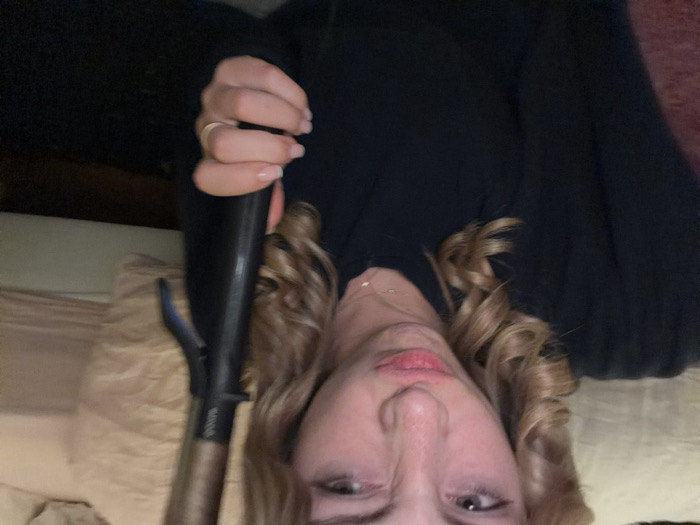 A young female, appearing to be in an indoor setting, is captured in a close-up, slightly blurred image. She has light skin, blonde hair styled in loose curls, and is wearing a dark, long-sleeved top with a delicate gold-colored necklace visible. Her eyes are partially narrowed, and her lips are puckered in a pouting expression, directed towards the camera. Her right hand, with natural-colored fingernails and a plain gold band ring on the ring finger, holds a black and metallic brown hair curling iron. The background is out of focus, revealing a light-colored object, possibly a bed or sofa, to the right, and a darker surface below. The image provides no discernable information regarding a specific location within Paris, France.