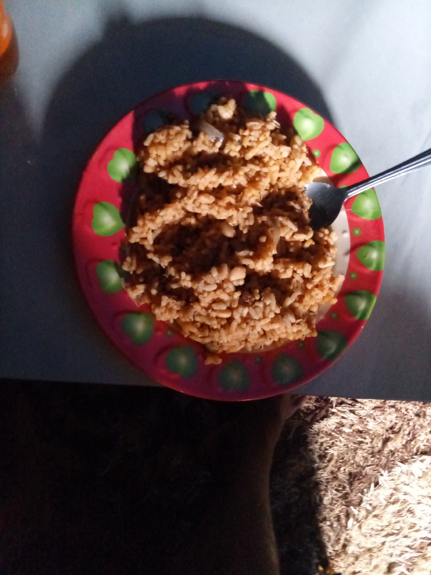 A top-down shot shows a red plate filled with brown rice, with a silver fork placed on top. The plate has a green heart pattern around the rim. The plate is sitting on a light-colored surface, possibly a table. A dark shadow is cast across the upper left of the image. The lower part of the image reveals a dark area and a patch of light-colored, textured material. The location is Unknown, Unknown.