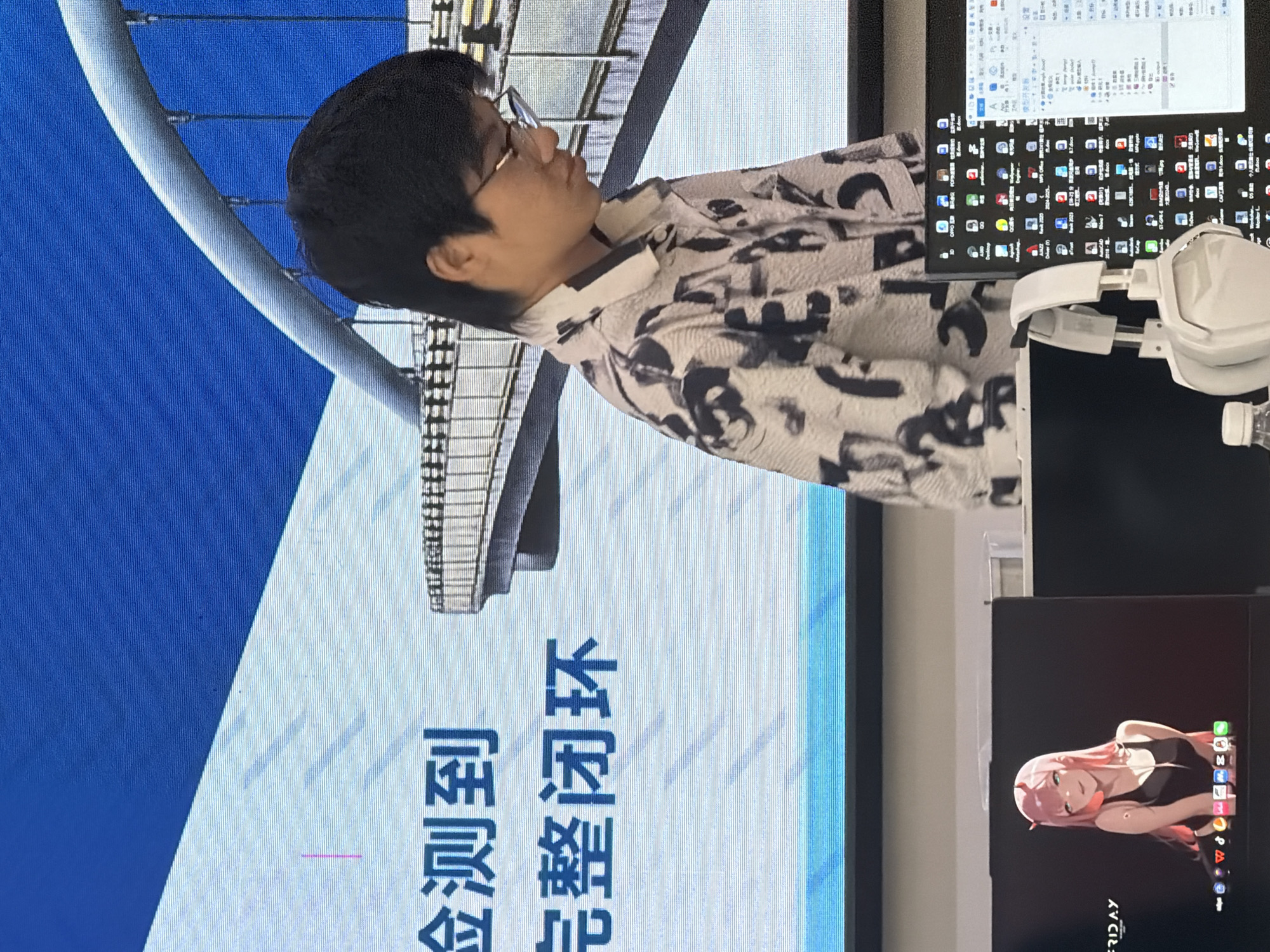 The image depicts an indoor scene, likely an office or a study, with a large screen in the background displaying a blue and white graphic with text. A young man wearing glasses and a black and white patterned shirt is standing in front of the screen. In the foreground, there are two computer monitors. The left monitor displays a desktop background of an anime character. The right monitor shows a computer interface with various icons and text. A pair of white headphones rests on the desk in front of the monitors, and a partially visible water bottle is also present. The overall lighting suggests an artificial indoor environment. The image does not contain any identifiable landmarks or clues that would allow for its location in Jiangjin, China, or any other specific city.