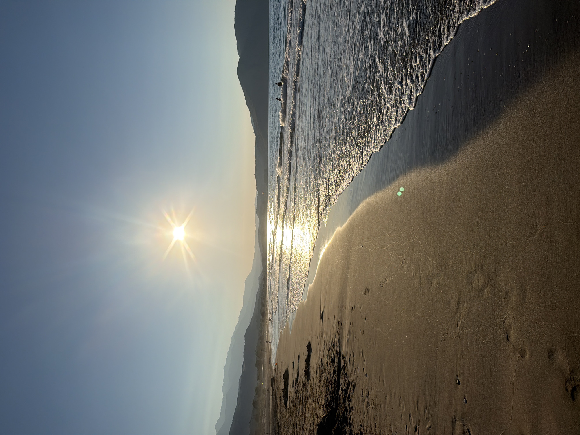 This image depicts a serene beach scene at sunset in Georgioupoli, Greece. The sun, positioned high in the clear blue sky, casts a warm, golden glow across the water and sand, creating a sparkling reflection on the sea. Gentle waves lap against the shore, leaving foamy trails on the wet sand. In the distance, silhouetted mountains form a hazy backdrop against the horizon.
The beach itself is sandy, with footprints visible in the foreground, indicating recent human activity. Some sandcastle structures are also present on the left side of the frame. A few individuals are visible in the water, appearing to be swimming or playing in the gentle surf. The overall atmosphere is peaceful and idyllic, capturing the beauty of a Grecian sunset by the sea.