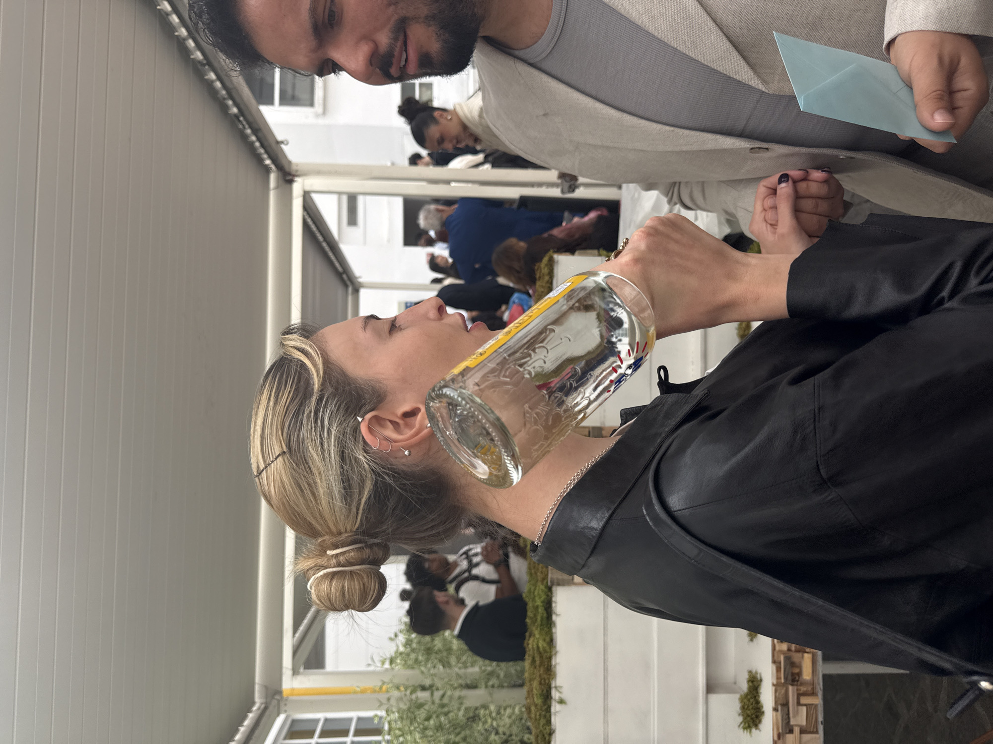 A woman is drinking from a glass bottle in an outdoor setting that appears to be a gathering of people. She has blonde hair styled in a bun and is wearing a black jacket. Her head is tilted back to drink from the bottle. A man in a light-colored suit is visible in the background and appears to be holding a blue envelope. Other people are visible in the background. The setting includes what looks like a patio area with plants and white architectural elements. Based on the visible architectural style, the scene could be located in Milan, Italy.