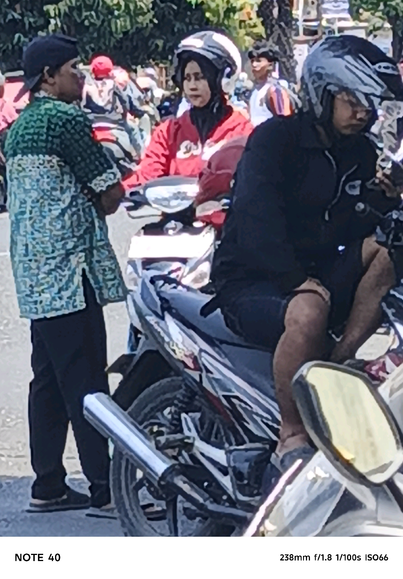 In Cilacap, Indonesia, two individuals are positioned on or near motorcycles. The person on the left, wearing a green patterned shirt and dark pants, faces right, looking towards the person on the right. The person on the right, wearing a black jacket and dark shorts, is seated on a motorcycle and looking downwards. Both individuals are wearing helmets. The environment appears to be outdoors, with trees visible in the background. The overall scene suggests a casual interaction or moment of rest.