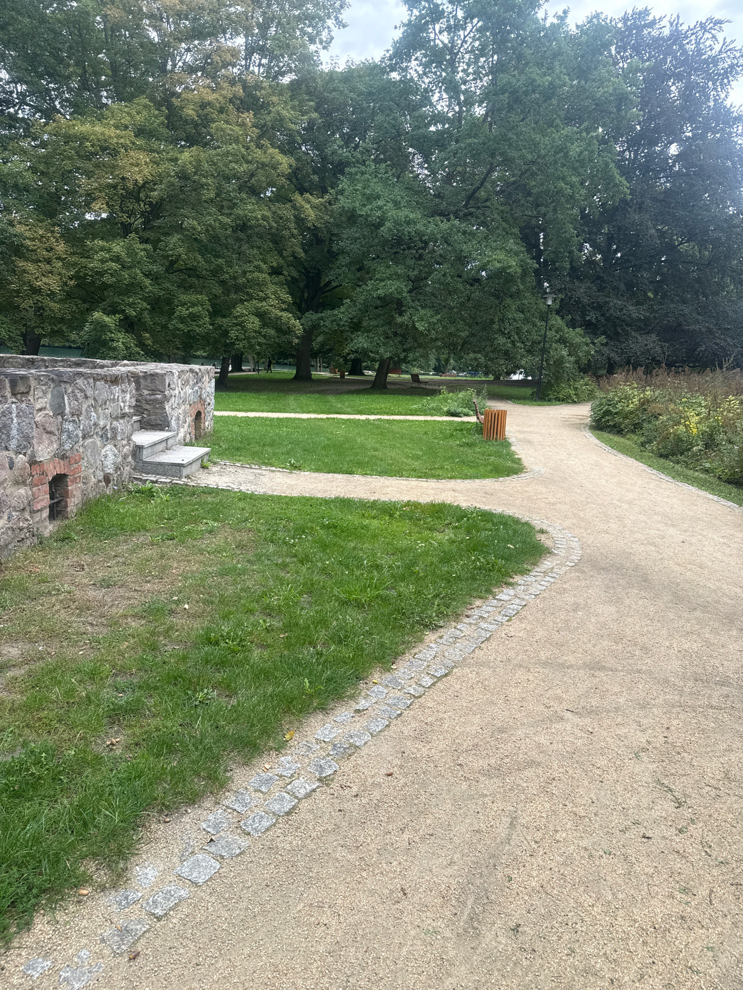 This is a photograph of a park path in Łagów, Poland. The path is made of light brown gravel and is bordered by small, grey stones. The path curves to the right. To the left of the path is a patch of green grass. Beyond the grass is a stone wall with two small arched openings. There are some stone steps leading up the wall. In the background, there are large, mature trees with green foliage. Some people can be seen walking in the distance. There is a wooden trash can near the path. The sky is overcast.
