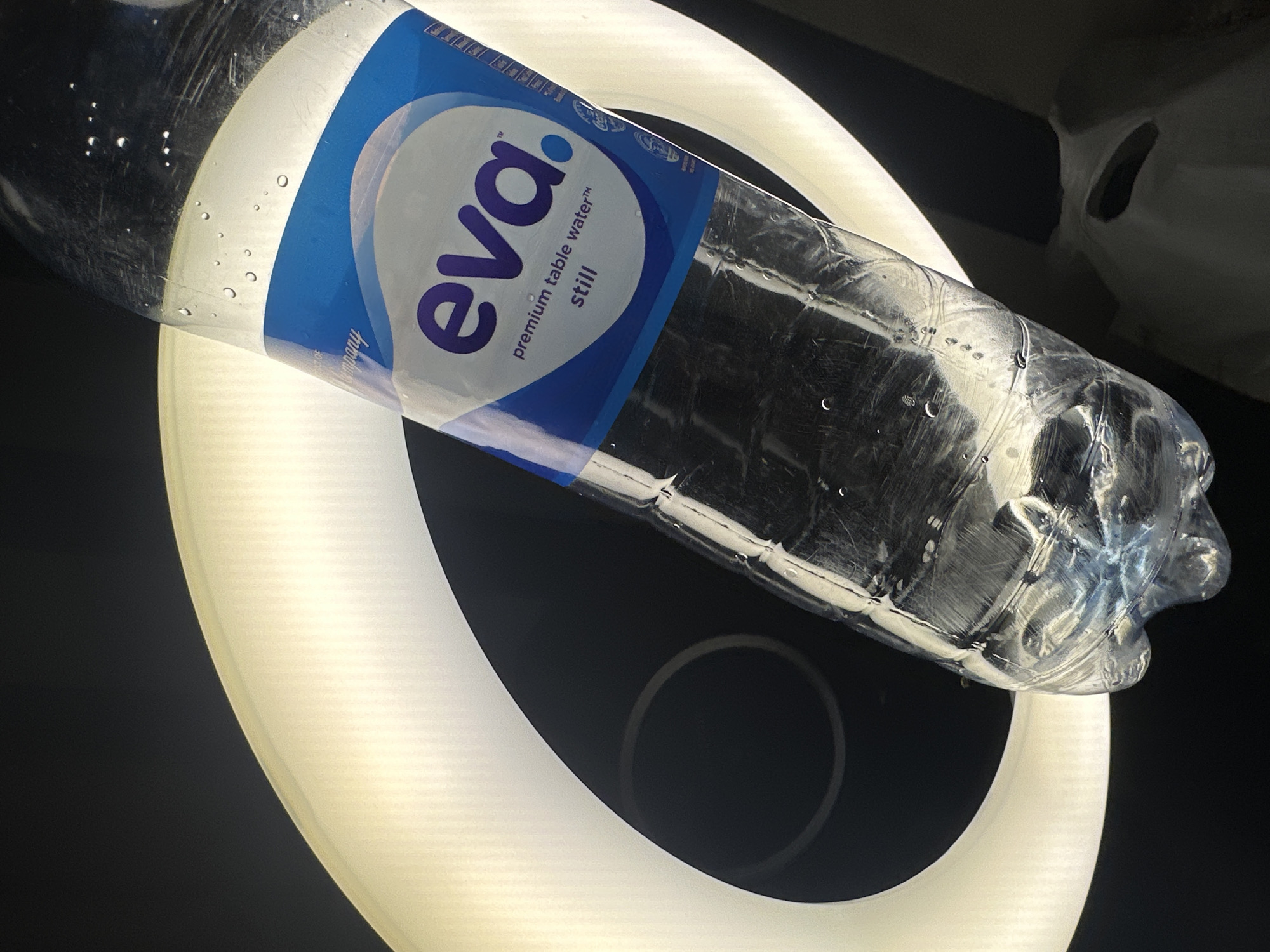 A plastic bottle labeled "eva. premium table water™ still" is positioned diagonally over a circular light. The background is black, and a blurry, white object is partially visible in the upper right corner. The scene is artificially lit with strong backlighting. The location of Orie Uratta, Nigeria is not evident within the image.