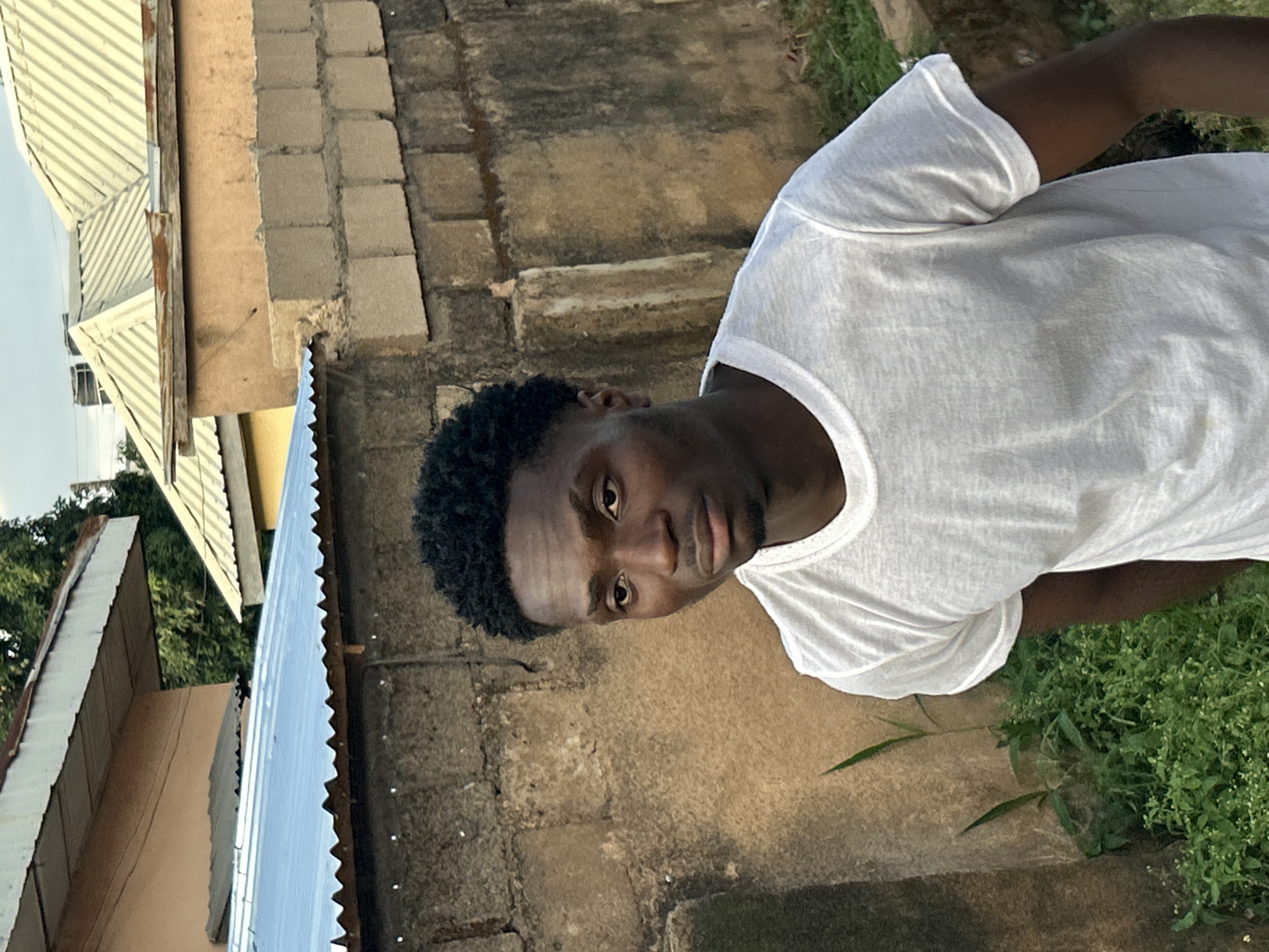 The image shows a young Black man, rotated 90 degrees clockwise, looking directly at the viewer with a neutral expression. He has short, dark, curly hair and is wearing a white short-sleeved t-shirt. His head is positioned towards the left of the frame, with his body extending diagonally across the image.
The background, also rotated, features various structures and natural elements. Directly behind the man is a textured wall made of concrete blocks or bricks, showing signs of wear. To the upper left, parts of buildings with distinctive corrugated metal roofs are visible, some light-colored and others with a blue-gray sheen. Green foliage and trees are also discernible in the distance behind these structures. In the lower right portion of the image, near the man's arm, a patch of vibrant green grass and small plants grows on the ground.
The lighting suggests it is daytime, with soft, natural light illuminating the scene and creating subtle shadows that highlight textures on the wall and the man's face. The man is posing for the camera, and no other specific activity is taking place. The scene depicts an outdoor setting, likely a residential or semi-urban area. The location context provided indicates this is in Jos, Nigeria. There is no visible text in the image.