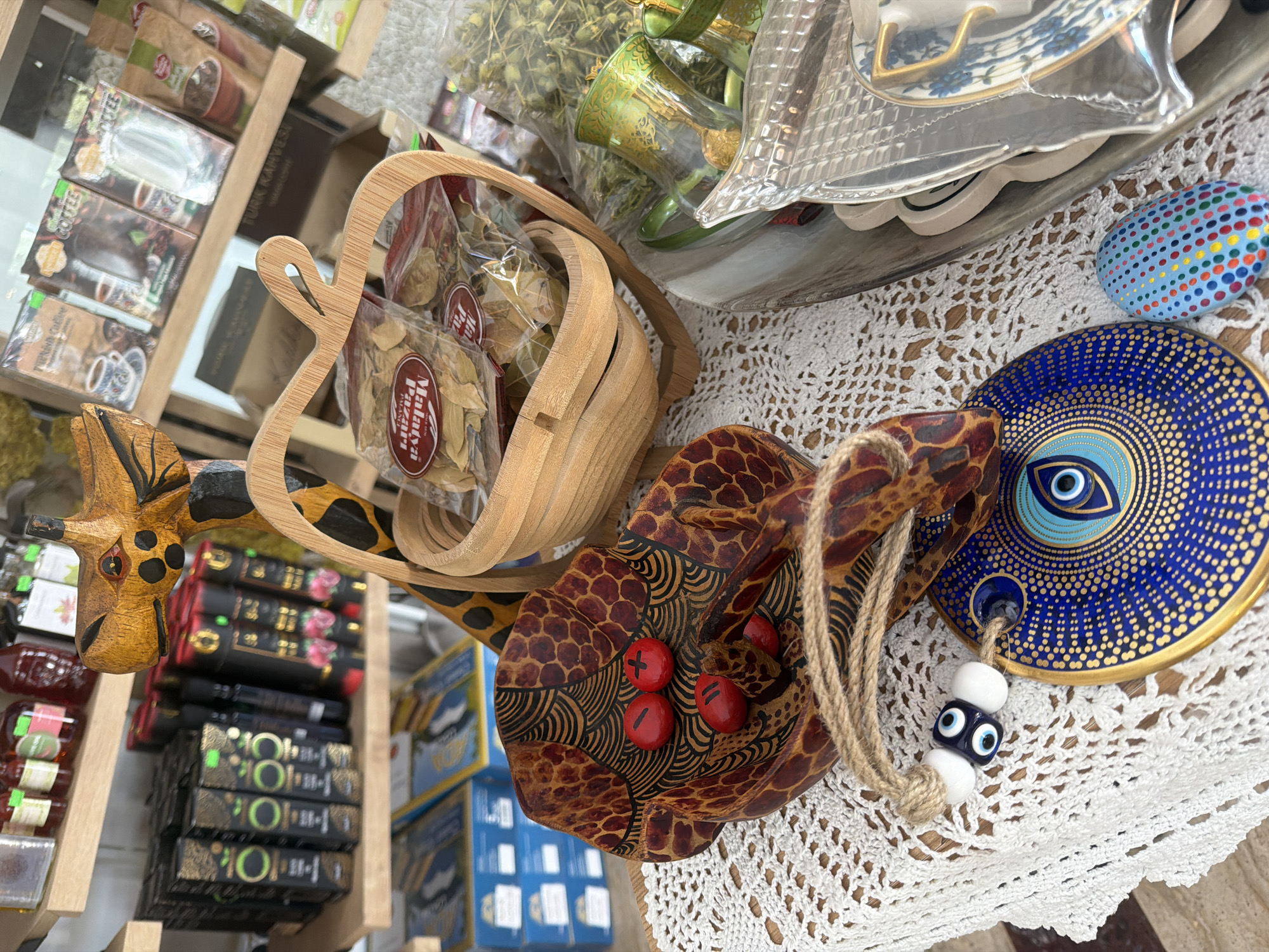 The image depicts a close-up, high-angle view of a retail display featuring various decorative and food items. In the foreground, a wooden carved giraffe figure, with a textured brown and black pattern, stands partially visible. A woven apple-shaped basket, also made of wood, rests in front of the giraffe. Inside the basket are several plastic-wrapped packages of dried herbs or spices. Below the basket and to the left, a decorative, dark brown, patterned bowl, likely ceramic or wood, contains three small red objects with markings resembling numbers or symbols, possibly dice or decorative stones. A rough rope hangs from the bowl, adorned with two small white beads and a blue evil eye charm with white and blue eyes. To the right of the bowl and further down is a round, blue, decorative plate with intricate gold patterns, also featuring an evil eye design. A small, oval-shaped, blue stone with colorful polka dots is placed next to the plate. The items are arranged on a white, lace doily.
In the background, wooden shelves are visible, stocked with various products. The top shelf holds several bottles of what appear to be jams or sauces. The middle shelf displays numerous black boxes with gold lettering and designs, and some cylindrical red packages with black and gold accents. The bottom shelf contains blue and yellow boxes. Further back and to the right, a collection of small green glass items with ornate gold patterns are visible, possibly tea glasses or shot glasses. The overall impression is of a shop selling artisanal goods, local crafts, and possibly food products, consistent with a souvenir shop or a specialty market.
The scene is illuminated by bright, natural light, suggesting an indoor setting with good lighting. The focus is on the meticulously arranged merchandise, creating an inviting and visually appealing display. Given the presence of what appear to be Turkish coffee packaging and artisanal goods, the location could be a shop in Marmaris, Turkey, a popular tourist destination known for its markets and souvenir shops. However, without explicit visual cues indicating Marmaris specifically (such as street signs, landmarks, or distinct architectural styles), this remains an inference based on the typical offerings in such locations.