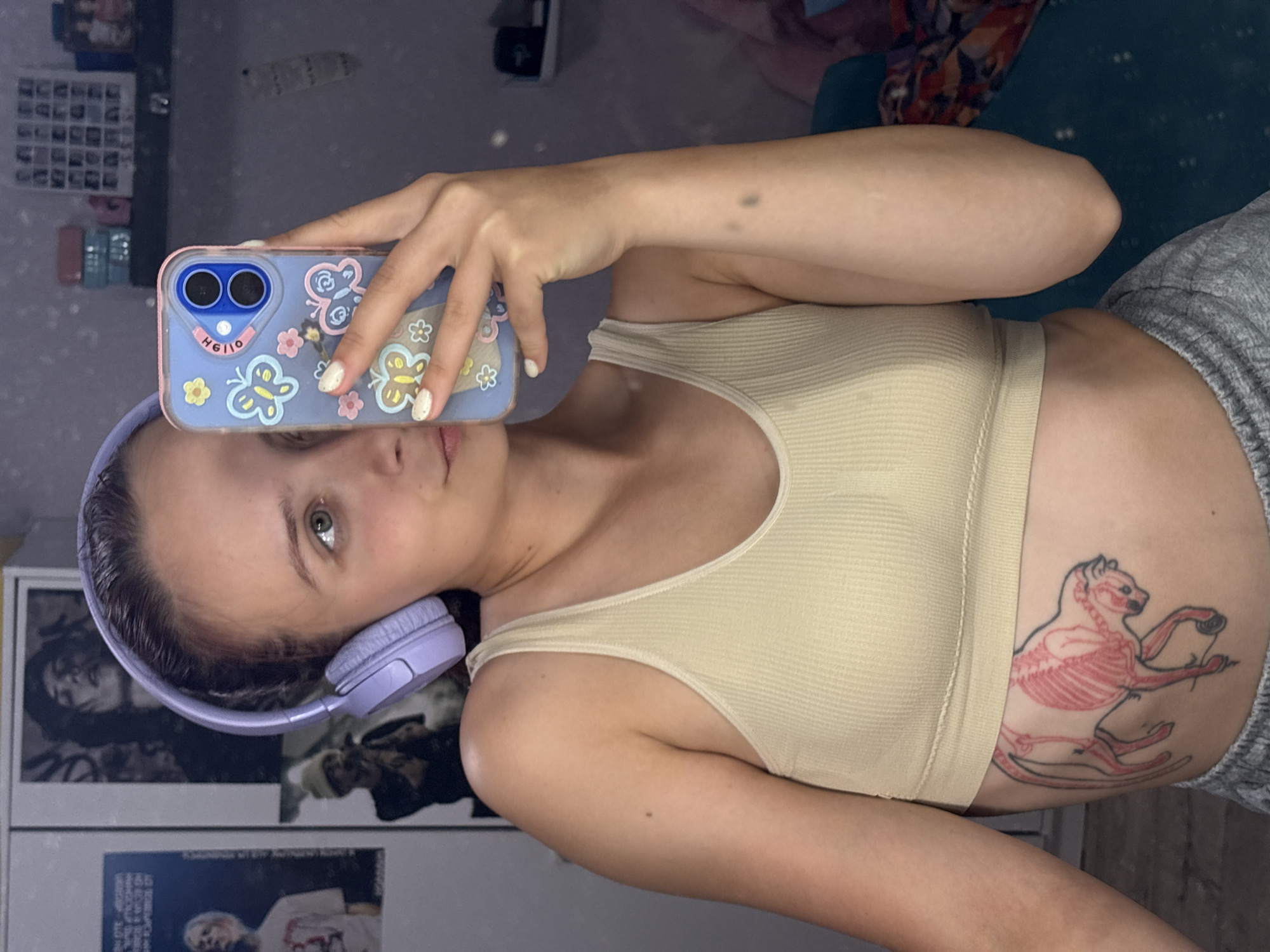 A young woman with light skin, dark hair, and visible green eyes is depicted in a mirror selfie, located in the city of Прикамский, Russia. She is wearing light purple over-ear headphones and a light yellow/beige ribbed crop top, with grey sweatpants partially visible. Her left hand holds a light blue smartphone encased in a clear cover adorned with multiple colorful butterfly and flower decals, obscuring the lower left portion of her face. The phone's dual camera lenses are visible, along with the word "HELLO" printed upside down on the case. Her visible fingernails are painted white with a speckled finish. A prominent tattoo on her left side, beneath her rib cage, depicts an animal resembling a cat or feline, with its skeletal structure (ribs, spine) rendered in red, outlined in black, appearing to be pushing a skateboard. A small, dark mole is present on her right upper arm, and two small, darker spots are on her right forearm.
The background, a reflection in the mirror, reveals a room with a light-colored wall or cabinet featuring several posters. One poster prominently displays a person with dark hair, another shows a person with light hair in a white t-shirt, and a third depicts a person wearing a beanie and holding a microphone. To the right, a dark shelf holds an array of small, framed photographs or decorative items arranged in a grid pattern, alongside stacked pink and blue containers. A blurred, dark teal object, possibly furniture, is visible on the far right.