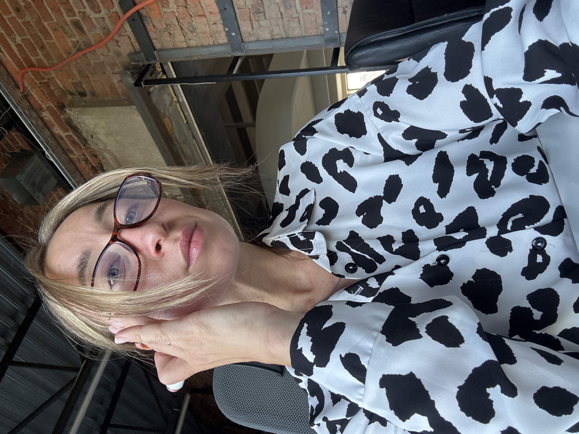 A woman with blonde hair and glasses is sitting in a chair, looking at the camera. She is wearing a black and white cow-print shirt. Her hand is raised to her ear. The background appears to be an industrial or office space with exposed brick and metal beams. The image was taken in the city of Chaikovsky, Russia.