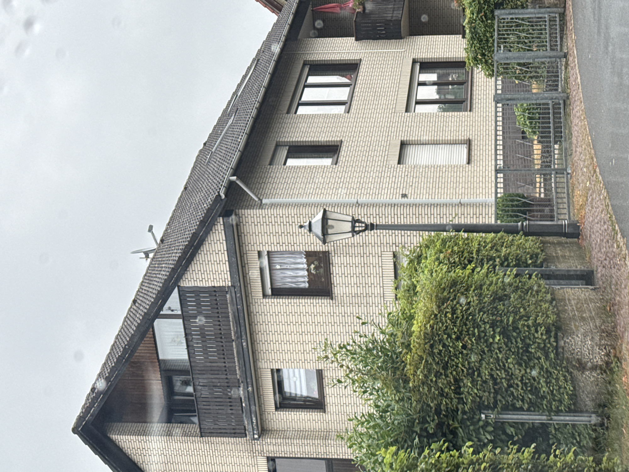 The image depicts the exterior of a residential building in Bad Sachsa, Germany. The building is constructed of light-colored brick and features a dark-tiled, pitched roof. There are multiple windows visible, some with brown frames and white roller shutters. A balcony with a dark wooden railing is present on the upper level. A black, ornate street lamp stands in front of the building. Lush green bushes and trees partially obscure the lower section of the building and a gray metal fence with decorative elements runs along the front. The sky is overcast, suggesting a cloudy day. The scene appears to be a quiet residential street.