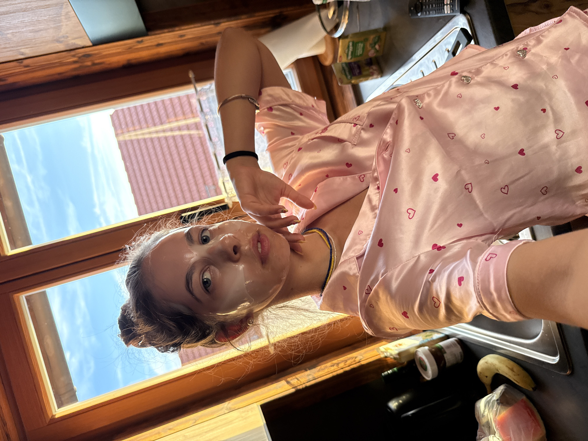 A young woman with her hair in a bun is taking a selfie in what appears to be a kitchen. She is wearing a pink satin pajama top with small red hearts printed on it and a face mask. She is leaning her head on her hand and her eyes are looking at the camera. The background shows a window with a view of the sky and some rooftops, as well as a kitchen counter with various items on it, including a banana, a jar of spread, and some packets. The scene is lit by natural light coming from the window. The location is not explicitly mentioned in the image or context.