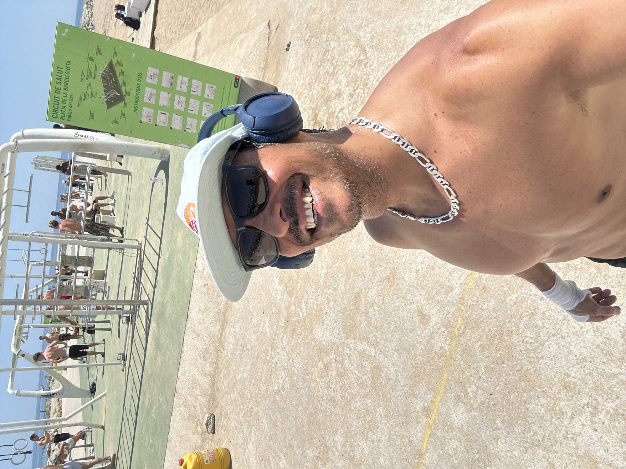 The image depicts an outdoor fitness area at Platja de la Barceloneta in Barcelona, Spain. The foreground features a shirtless man wearing a light blue cap, sunglasses, and blue headphones, with a silver necklace and a white bandage on his left wrist. He is smiling and looking towards the camera, appearing to take a selfie. Behind him, a green sign displays "CIRCUIT DE SALUT PLATJA DE LA BARCELONETA Espigó del Gas" and "INSTRUCCIONS D'ÚS" with diagrams of exercises. The background shows various exercise equipment, including pull-up bars and monkey bars, with several other individuals engaged in workouts or resting. The ground is a mix of sand and paved concrete, with the Mediterranean Sea visible in the distance. A yellow container is also visible on the left side of the image.