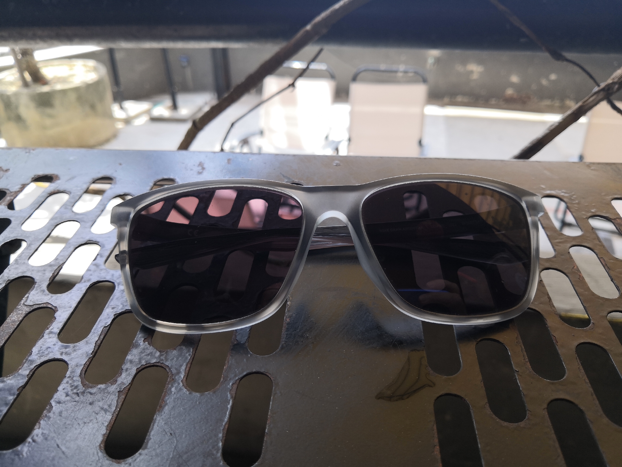 A pair of semi-transparent gray framed sunglasses with dark lenses is resting on a perforated metal surface. The surface appears to be a table or bench with elongated oval cutouts. In the background, a blurry scene shows light-colored structures, possibly furniture or partitions, with some greenery and branches visible overhead. The lighting suggests an outdoor setting, possibly a balcony or patio area. The specific location in Bac Nha Trang Ward, Vietnam is not discernible from the visual information provided in the image.