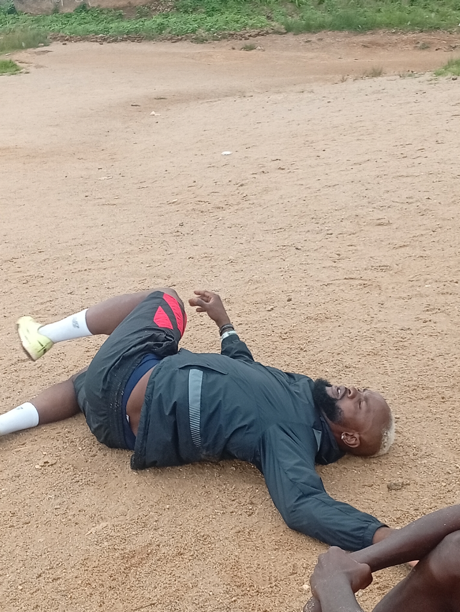 A person with blonde hair and a beard, wearing a black jacket and grey shorts with red accents, lies on their back on a dirt field. Their right leg is bent at the knee, with their foot extended. Their left leg is bent at the hip and knee, with their foot on the ground. Their right arm is bent at the elbow, with their hand near their head. Their left arm is extended along their body. Another person's arm is visible in the lower right corner, holding the right arm of the person on the ground. The background shows a dirt field with some scattered debris and green foliage in the distance, suggesting an outdoor environment, possibly in Jos, Nigeria.