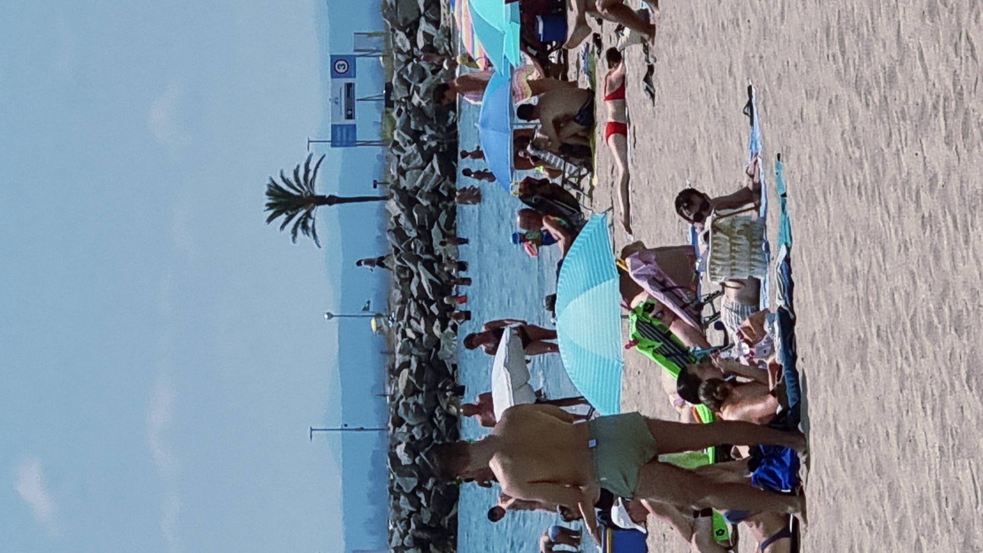 The image depicts a crowded beach scene in Castelló d'Empúries, Spain, likely during a sunny day with a clear sky. The foreground is dominated by sand, with several people sunbathing and relaxing. A man in light green shorts stands in the left foreground, facing away from the camera. To his right, a woman is lying down, partially obscured by a beach umbrella. Further back, multiple individuals are seen under beach umbrellas or lying on towels. Some people are wading in the ocean, which appears calm and blue. In the middle ground, a breakwater made of large rocks separates the beach from the sea. Beyond the breakwater, a palm tree is visible, along with distant hills and some buildings. A sign with a "3" is also present in the background. The overall atmosphere is one of leisure and recreation on a summer day.