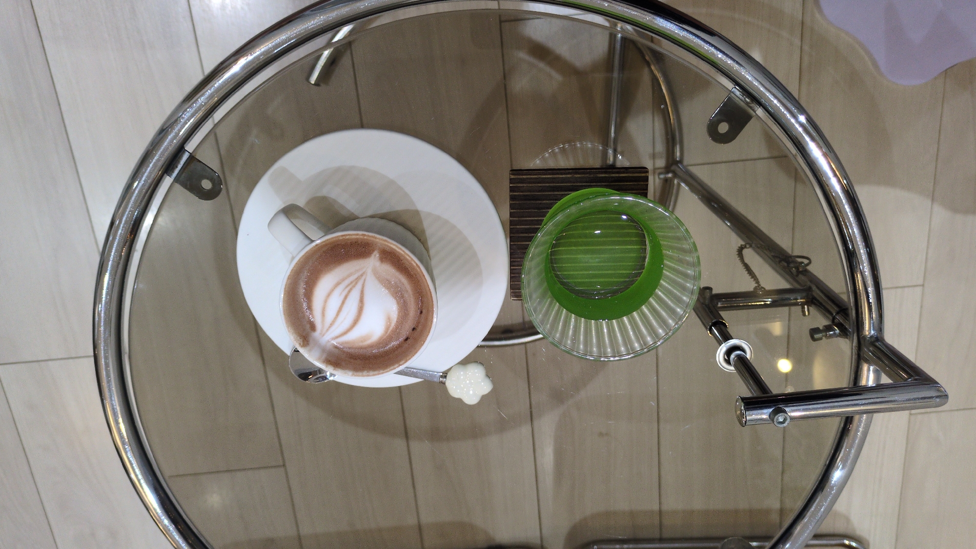 A high-angle shot shows a glass-topped table with a chrome frame, likely in a cafe or similar setting in Phan Rang – Tháp Chàm, Vietnam. On the table is a white cup and saucer containing a cappuccino or latte with latte art, accompanied by a small spoon with a flower-shaped embellishment. Next to the coffee, there's a small, transparent glass bowl with a green rim resting on a dark brown coaster. The background shows a neutral-toned floor, possibly tile or wood laminate, and a hint of a white or light-colored wall. No people are visible.