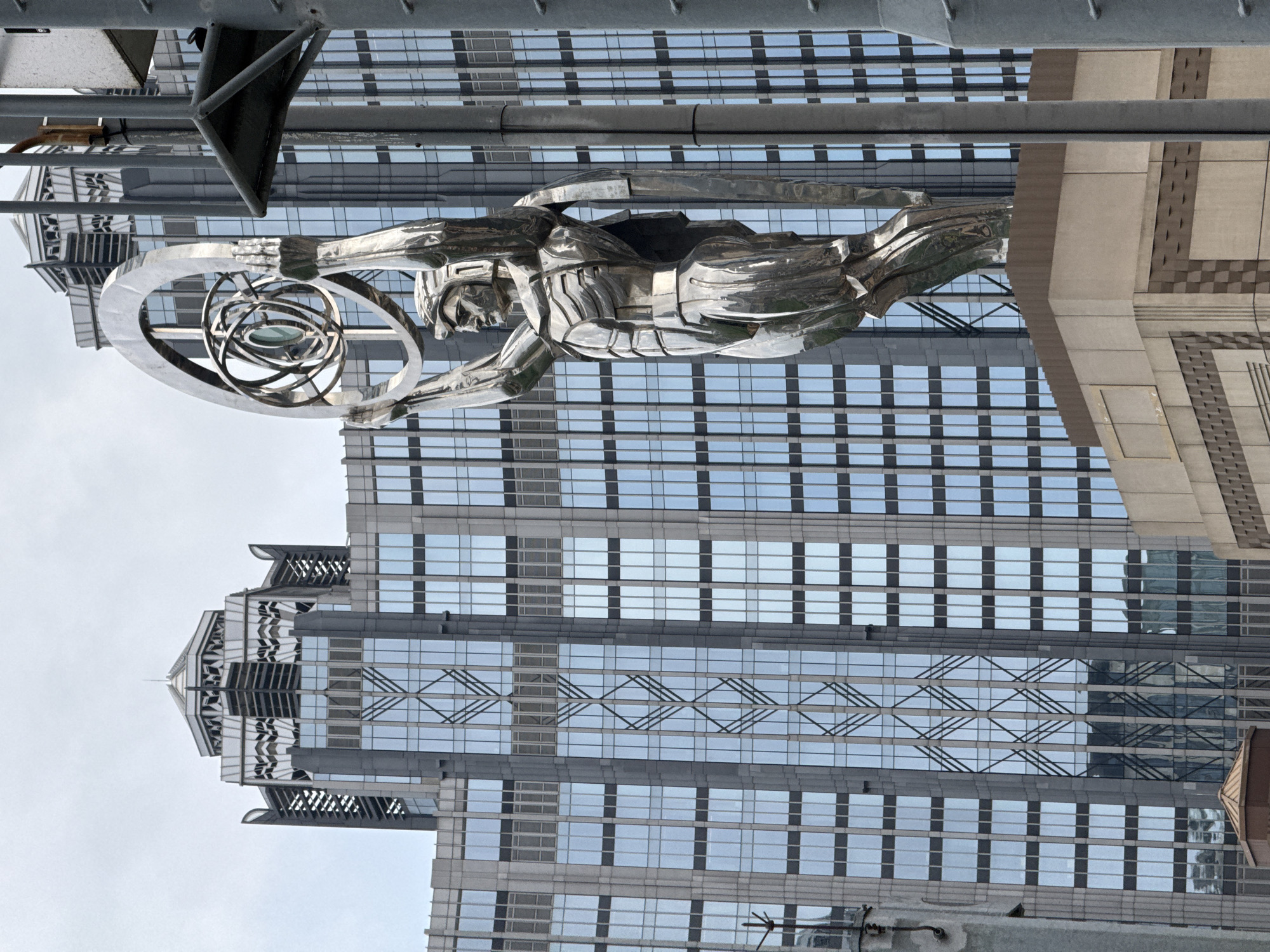 The image shows a large, chrome statue of a person holding a circular device with concentric rings in front of a tall skyscraper. The building has a distinctive stepped design at the top and a patterned facade of alternating dark and light glass. A section of metal framework is visible at the top of the image, partially obscuring the statue. The location is Unknown, Macao.