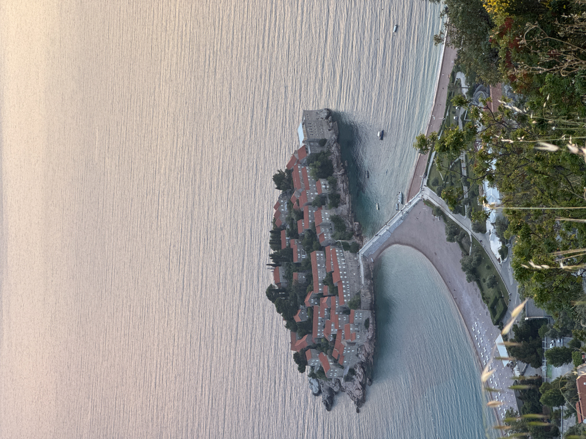 A high-angle view shows the island of Sveti Stefan, Montenegro. The island is connected to the mainland by a narrow isthmus of land. Red-roofed buildings are visible on the island, surrounded by trees. The water surrounding the island is a pale blue color. The coastline on the mainland features a beach with white umbrellas. Green trees and shrubbery are also present on the mainland. The photograph appears to be taken from a vantage point on a hillside or elevated position.