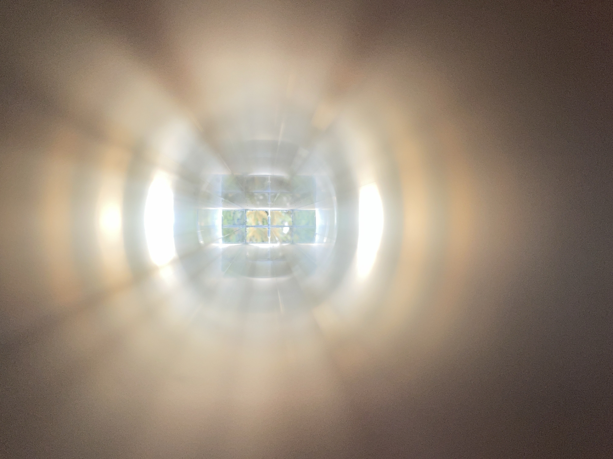 The image shows a view through a window, with a blurry effect radiating outwards. The window appears to be divided into multiple panes, offering a glimpse of greenery outside, possibly trees. The surrounding wall is a neutral, light tan color. The light is very bright around the window, creating a strong contrast with the dimmer areas of the wall. Without additional information, I can't identify a specific location in Burgas, Bulgaria.