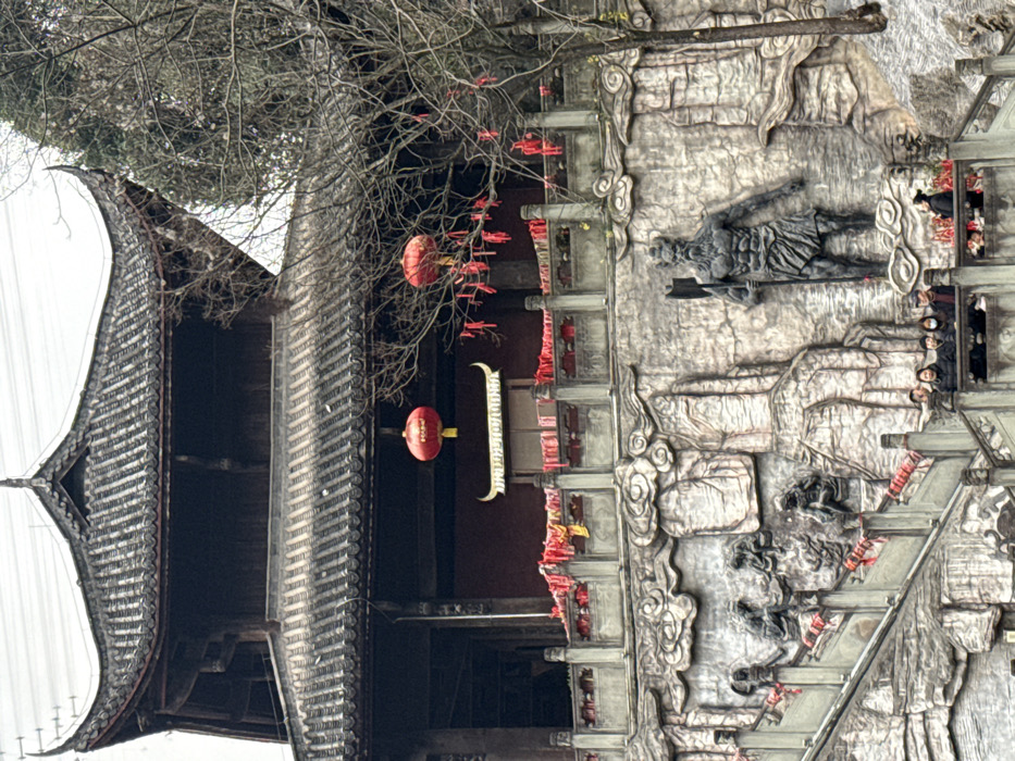 This image depicts a multi-level traditional Chinese architectural complex situated on a hillside in Pengshui Miao and Tujia Autonomous County, China, under an overcast sky.
The upper portion of the scene features a dark, two-tiered building with traditional curved, tiled roofs. Its walls appear to be constructed of dark wood. A balcony on the second level is adorned with numerous red fabric strips tied to its balustrade. Two circular red Chinese lanterns hang from the eaves of the second story, and a gilded, horn-shaped decorative element is visible above an opening on the same level.
Below the main building, a grey stone wall is intricately carved in high relief, depicting stylized rock formations and clouds. Prominently featured on the right side of this carved wall is a large, dark grey relief sculpture of a muscular, bearded figure with a horned headpiece, wielding an axe. To the left of this figure, smaller human-like figures are sculpted into the rock formations, appearing to be climbing or interacting with the environment.
A wide grey stone staircase with balustrades ascends diagonally from the lower left towards the architectural complex. Red fabric strips are tied to the balustrades of this staircase. On a mid-level landing of the staircase, a group of approximately 6-8 individuals, dressed in dark winter clothing, are gathered, observing the structures above. They include men and women, some wearing face masks. Bare branches of a deciduous tree, also adorned with red fabric strips, partially frame the right side of the image.
