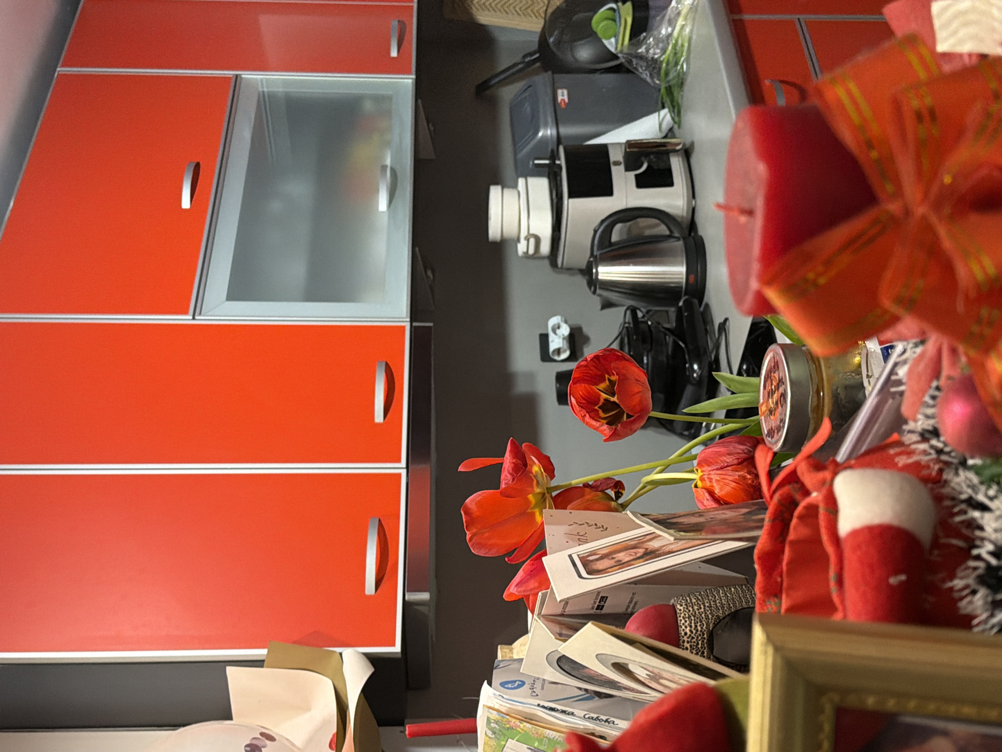 A close-up, slightly angled, indoor shot displays a cluttered surface in Varna, Bulgaria. On the left side of the frame stands a red cabinet with horizontal silver handles, partially obscuring the grey wall behind the objects. A stack of items including tulips, cards with faces, a small jar with a label, Christmas decorations (including a red and white stocking and a red bauble), and a gold picture frame are placed towards the right of the image. Kitchen appliances such as a mixer, a kettle, and a plastic container, are also visible. Overall, the image captures a festive and somewhat untidy domestic setting.