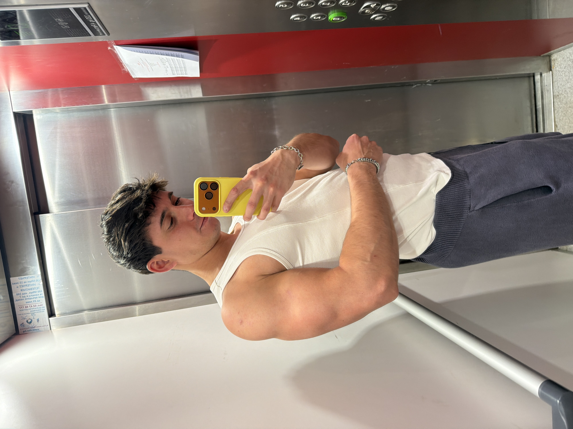 A young man takes a mirror selfie inside a modern elevator in Sofia, Bulgaria. He holds a bright yellow smartphone with a triple-lens camera module, positioned to capture his reflection. He is wearing a white ribbed tank top, revealing muscular arms, each adorned with a silver chain bracelet. Dark blue or grey sweatpants complete his casual attire.
The elevator interior features polished stainless steel panels and a prominent red horizontal accent strip. Above the man's reflected head, an elevator control panel displays several grey buttons and one illuminated green button. On the red panel, a document in a clear protective sleeve contains visible text, partially obscured and mirrored. OCR analysis of this text reveals fragments such as "39 52 1689", "Танятии", "NHLEGHELH", "RENANT", "001 ден адам 001", and a web address "gil.nelv.www". To the man's left, a vertical information panel, also mirrored, displays Cyrillic text, including "ТАННЕТНЕН", "NNENZUU", "001 ДЕН АДАМ 001", and "pd.noiw.www", likely providing instructions or safety information. A white ledge or shelf is visible at the bottom of the reflected scene. The man's expression is focused as he captures the photo.