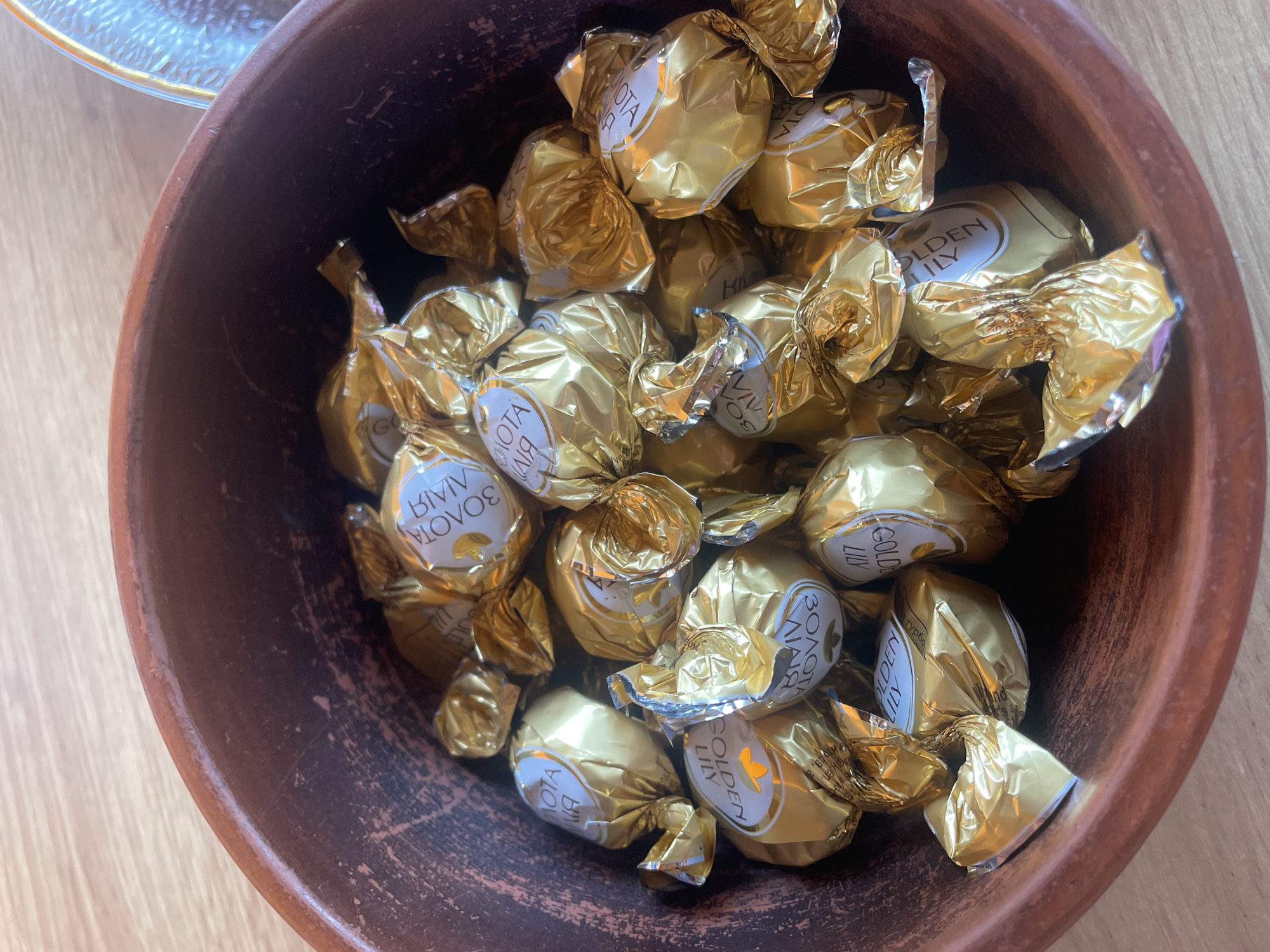 The image displays an overhead shot of a rustic, reddish-brown terracotta bowl filled with numerous individually wrapped chocolates. Each chocolate is encased in shiny golden foil. Visible on several wrappers are white labels bearing the text "GOLDEN LILY" in English and "ЗОЛОТА ЛІЛІЯ" in Cyrillic, indicating the brand "Golden Lily." The chocolates are densely packed within the bowl, with some wrappers appearing slightly crumpled. The bowl itself has an unglazed or roughly finished texture with visible imperfections and variations in its reddish-brown hue. The object is situated on a light-colored wooden surface, displaying distinct grain patterns. A portion of a clear, textured glass object, possibly another dish, is visible in the upper right quadrant of the frame. This scene is documented in Petrivka, Ukraine.