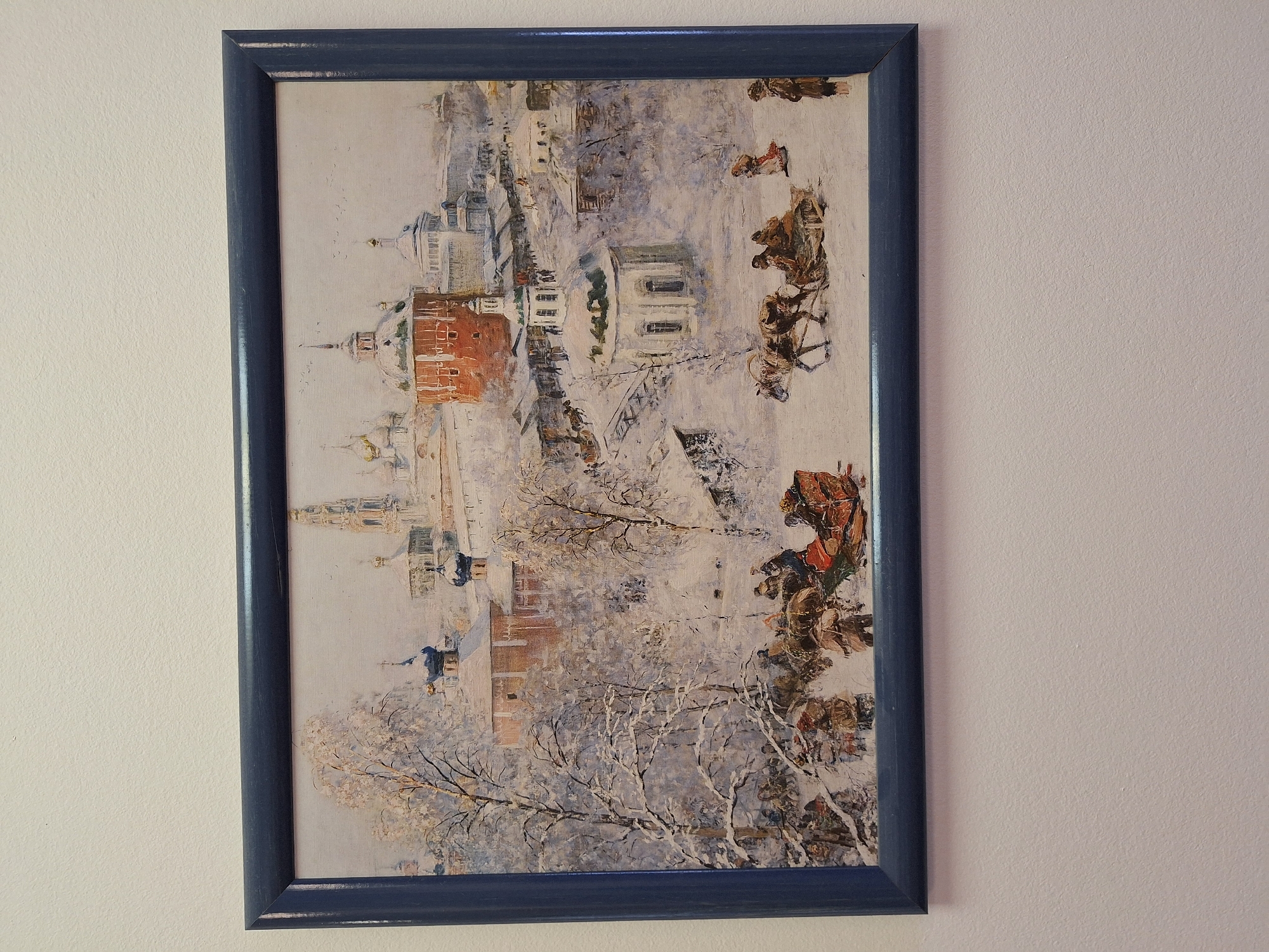 A framed painting depicting a bustling winter scene in Moscow, Russia, hangs on a light-colored wall. The artwork, characterized by an impressionistic style with visible brushstrokes, portrays a snow-covered cityscape under what appears to be a bright winter sky.
The central scene features several traditional Russian buildings, including structures with distinctive onion domes and spires, suggesting churches or cathedrals. Some buildings are rendered in brick-red hues, contrasting with the dominant whites and pale blues of the snow and sky. Bare trees, their branches laden with snow, are scattered throughout the landscape.
The primary activity depicted is movement along snow-covered paths or streets. Multiple horse-drawn sleighs and carriages, each carrying figures bundled in winter clothing, are visible, suggesting travel or daily commerce. People are driving the sleighs and walking alongside them.
Notable details include the heavy snowfall covering all surfaces, indicating cold winter weather. The lighting suggests it is daytime. The painting evokes a historical or traditional depiction of Moscow during winter.
The painting is enclosed in a dark blue or grey-blue wooden frame, which shows some signs of distressing or wear, adding to its character. No visible text appears within the painting or on the frame.