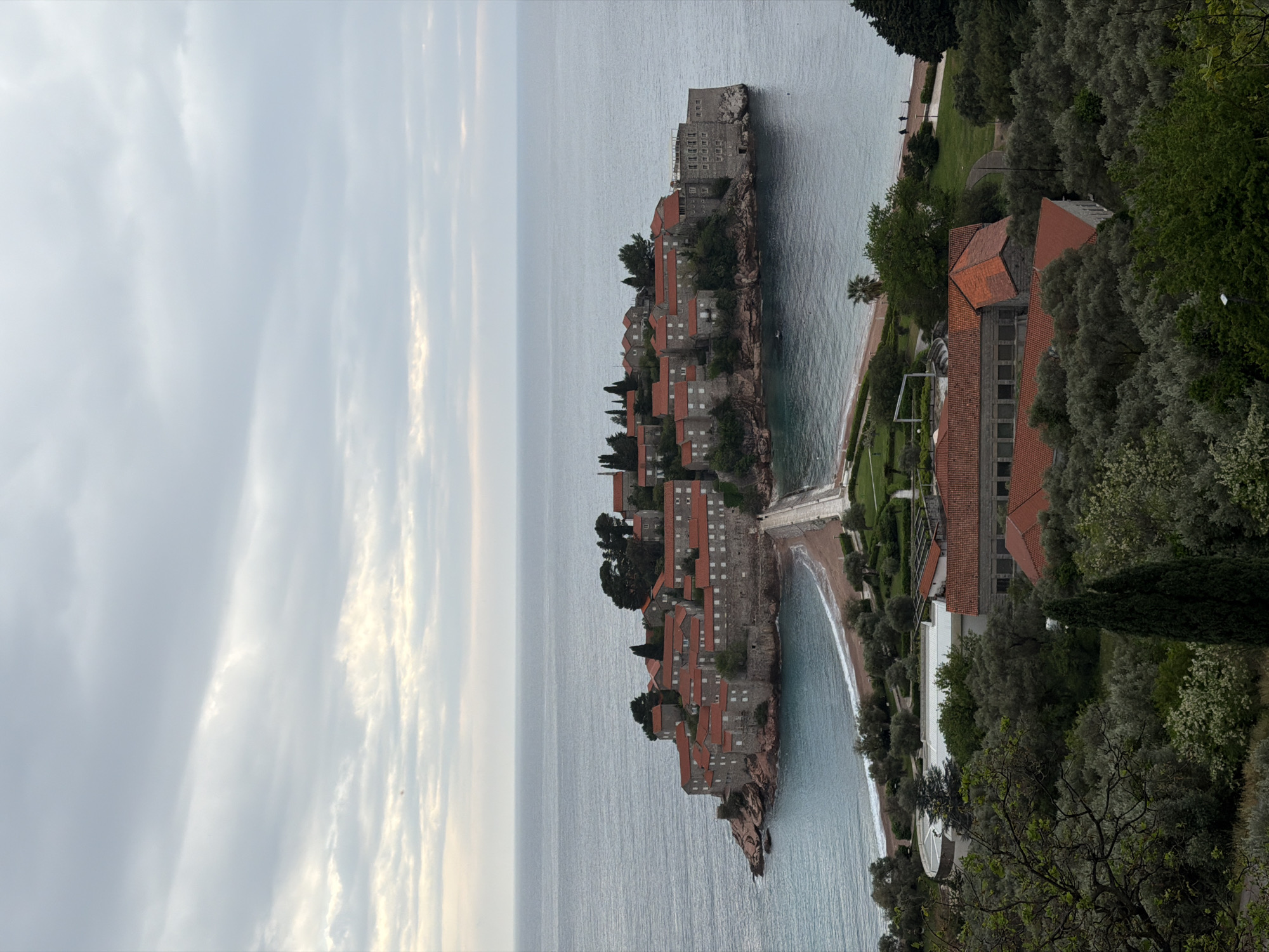 This image captures a panoramic view of Sveti Stefan, a picturesque fortified island village located in Montenegro. The scene is dominated by the unique architecture of the island, which is connected to the mainland by a narrow causeway.
On the island, numerous stone buildings with distinctive red-tiled roofs are densely packed, rising from the rocky outcrop. These structures appear historic and blend seamlessly with the natural landscape. The surrounding sea is calm, with a muted blue-gray tone, reflecting the soft light from the sky. On either side of the causeway, small beaches with light-colored sand or pebbles gently curve along the coastline.
The mainland portion of the image shows lush, verdant vegetation, including numerous trees and shrubs, indicative of a Mediterranean climate. Among the greenery, several buildings with red-tiled roofs are visible, likely part of a resort or private estate, nestled within well-maintained gardens. Pathways and manicured lawns can be discerned within the green areas.
The sky above is largely overcast with soft, diffused clouds, suggesting either an early morning, late afternoon, or a generally cloudy day. The light is gentle, enhancing the tranquil and serene atmosphere of the coastal landscape. No specific activities or people are visible in the frame, emphasizing the natural beauty and architectural grandeur of the location. There is no visible text in the image.