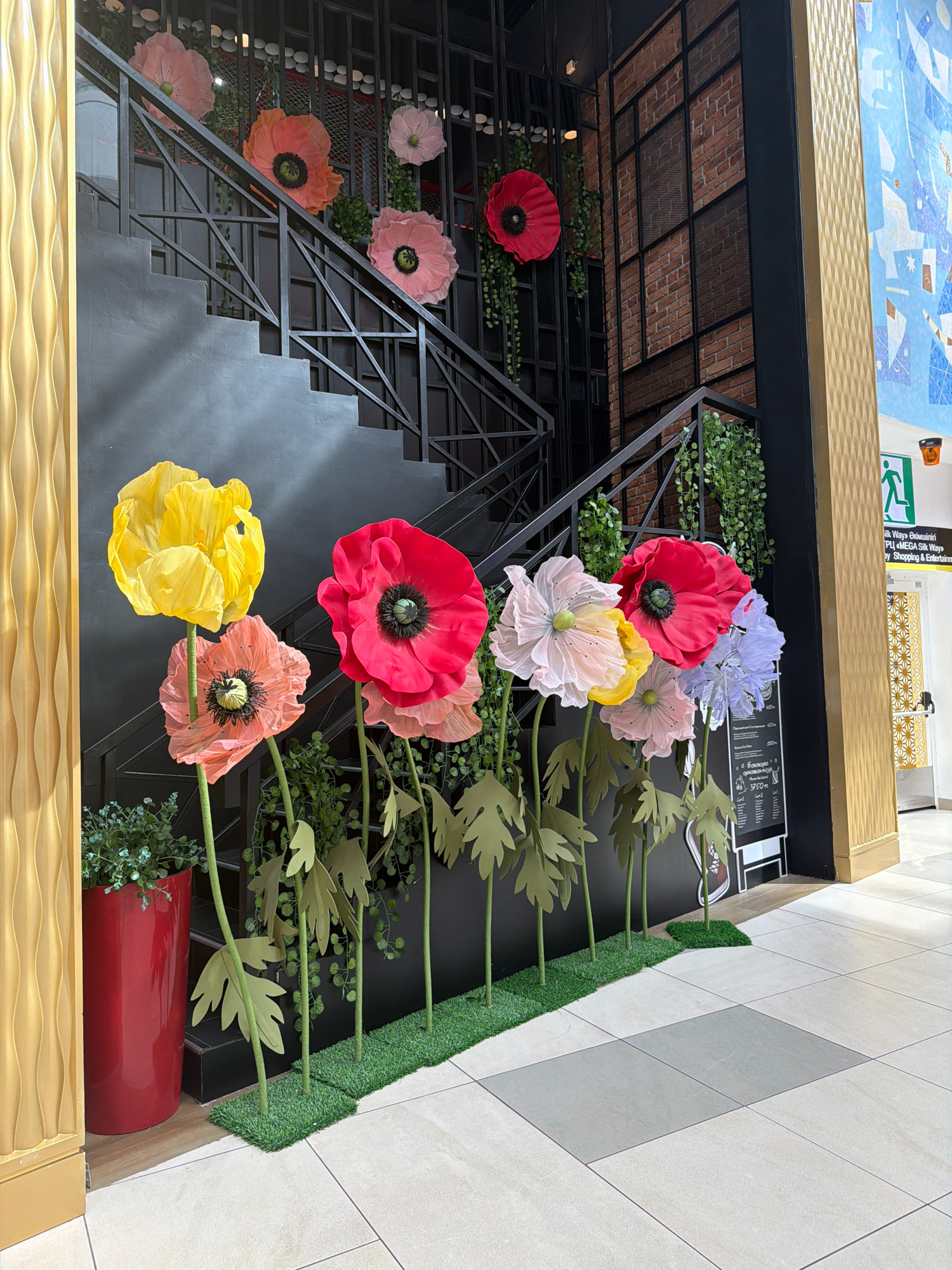 The image shows an indoor scene, likely a public space such as a mall or atrium, featuring a prominent staircase and decorative elements. The staircase is black metal with dark grey steps and a black railing. Along the railing and on the steps, large, colorful paper or fabric poppies are displayed as decoration. These flowers are in various shades, including yellow, pink, red, light pink, and light purple, with black centers. Some of the poppies are attached to the railing, while others are mounted on green stems with large green leaves, creating a garden-like effect at the base of the stairs. A bright red planter filled with greenery is positioned to the left of the stairs. In the background, to the right of the staircase, a brick wall with a black metal grid is visible. Further to the right, a gold-colored textured wall runs vertically, and a large abstract blue and white mural is partially visible. A sign with an arrow and text in English and Cyrillic is also present on the right side of the image, indicating "MEGA Silk Way | Shopping & Entertainment." This suggests the location is within the MEGA Silk Way shopping and entertainment complex in Astana, Kazakhstan. The floor is tiled with large light-colored tiles, with some grey tiles forming a contrasting pattern. The overall impression is a decorative installation designed to enhance the aesthetic appeal of the space.