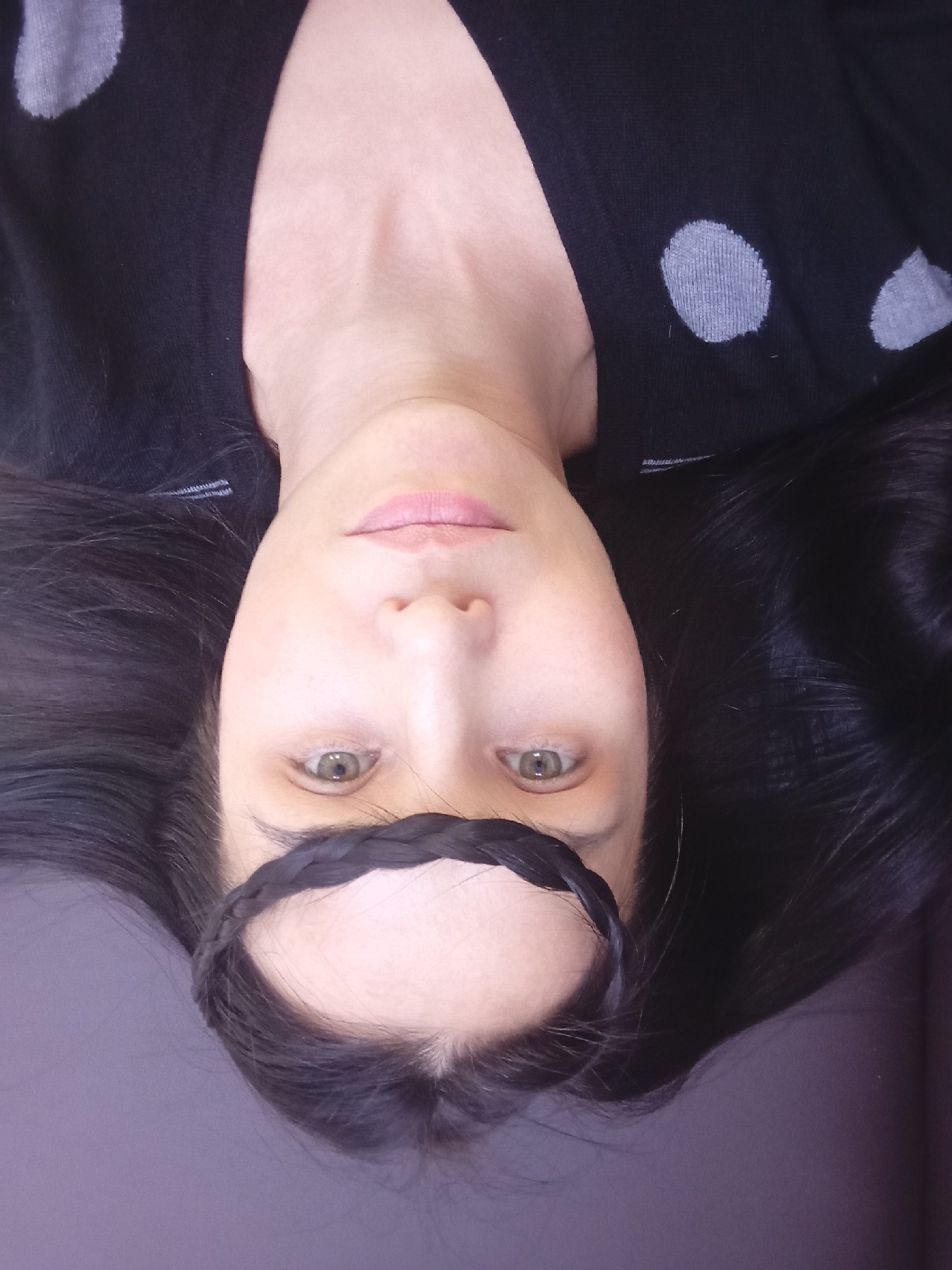 A close-up, upside-down portrait features a person with long, dark hair and fair skin. The person, who appears to be female, is lying down, with their head oriented so that the top of their head is at the bottom of the frame and their chin is towards the top. Their eyes, which are a greenish-hazel color, are open and directed towards the viewer. The lips are lightly colored and slightly parted.
A dark braid of hair is styled across the forehead, serving as a decorative element. The person is wearing a black top or sweater with light grey polka dots visible on the shoulders and upper chest area. The long, dark hair is spread out around the head, partially obscuring the background. The background appears to be a plain, dark purple or grey surface, possibly a bed or couch. No specific time of day or location cues are visible within the image itself, though the context provided is Izhevsk, Russia. No visible text is present.