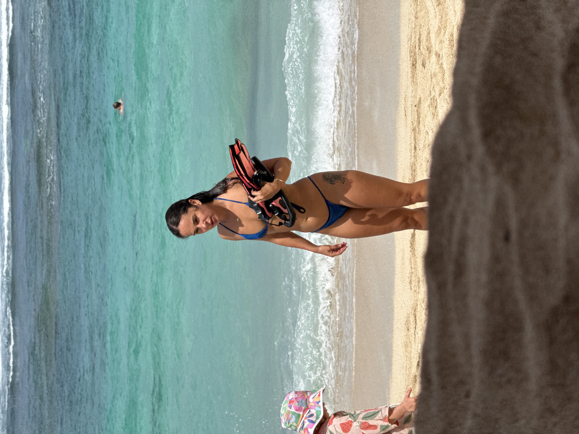 A woman in a blue bikini stands on a sandy beach next to turquoise water in Kuta Selatan, Indonesia. She is holding black and red diving fins. A person swims in the ocean behind her. In the lower foreground, a child with a floral hat and strawberry-patterned clothing is visible. Part of a large rock or cliff face is present on the right edge of the frame.