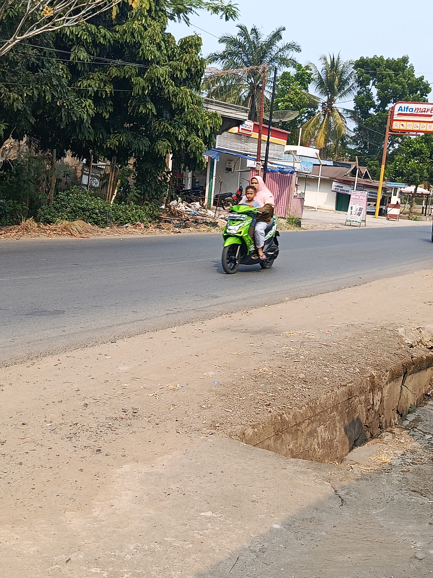 A medium shot captures a woman wearing a pink hijab and a light blue shirt, riding a bright green scooter with a child seated in front of her. The child, wearing a dark shirt, looks towards the camera. They are on a paved road with a sandy shoulder. In the background, palm trees and dense green foliage are visible, with a clear blue sky above. A sign for "Alfamart" is prominent on the right side of the frame, along with other storefronts and structures. The scene appears to be a street in the city of Talang Ubi, Indonesia.
