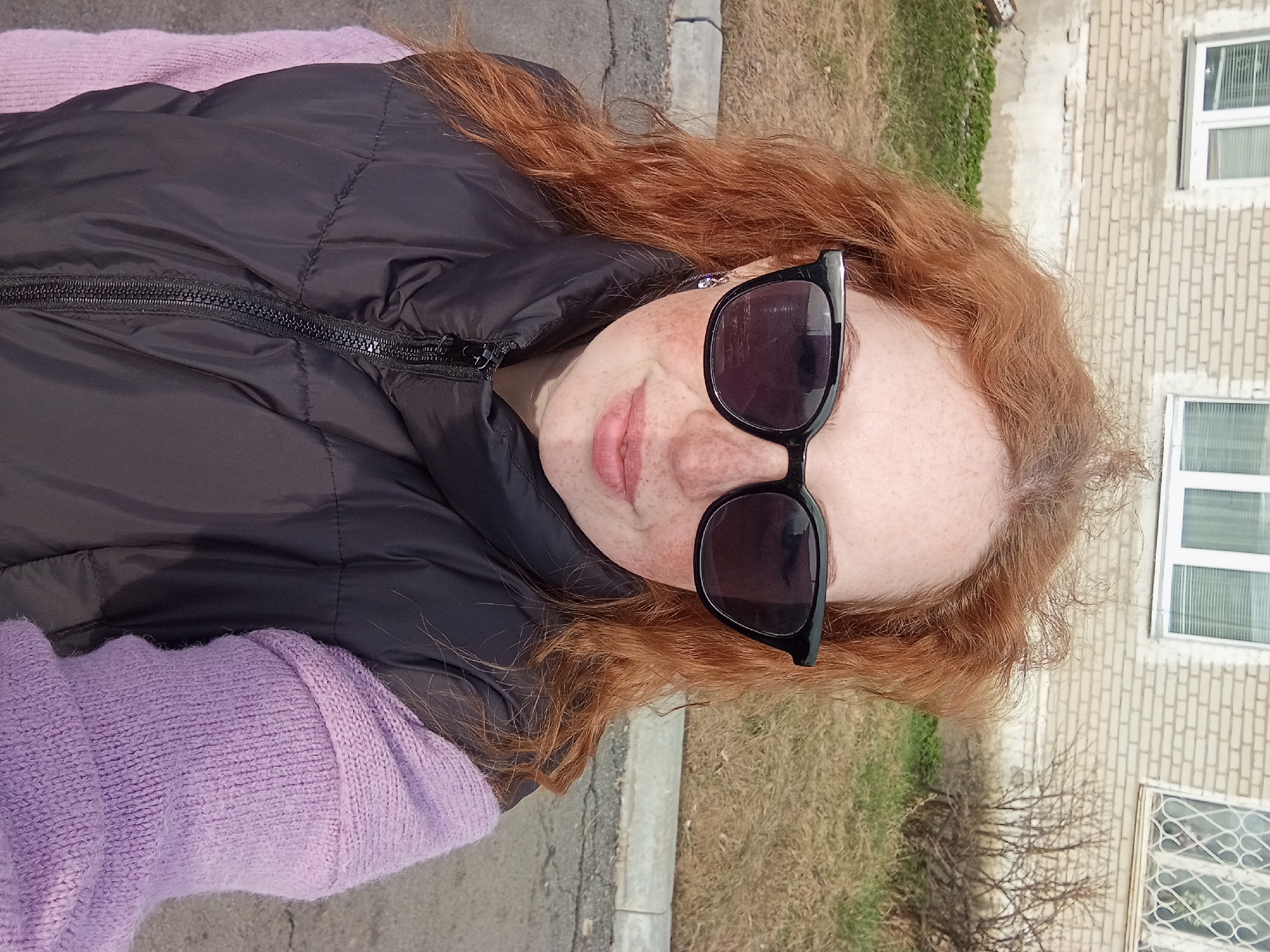 A woman with red hair and freckles is taking a selfie outdoors. She is wearing sunglasses, a black vest, and a lilac sweater. The background includes a brick building with windows, grass, and a sidewalk. The location is likely in Yurgamysh, Russia.